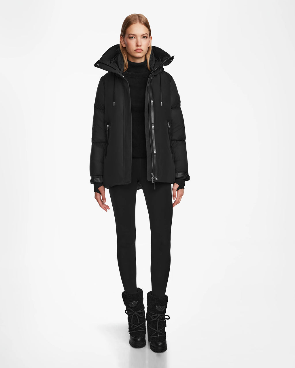 A woman faces forward against a plain white background, wearing the Rudsak LOLI - 8124507 Black womens down parka, paired with a black turtleneck, black leggings, and black winter boots.