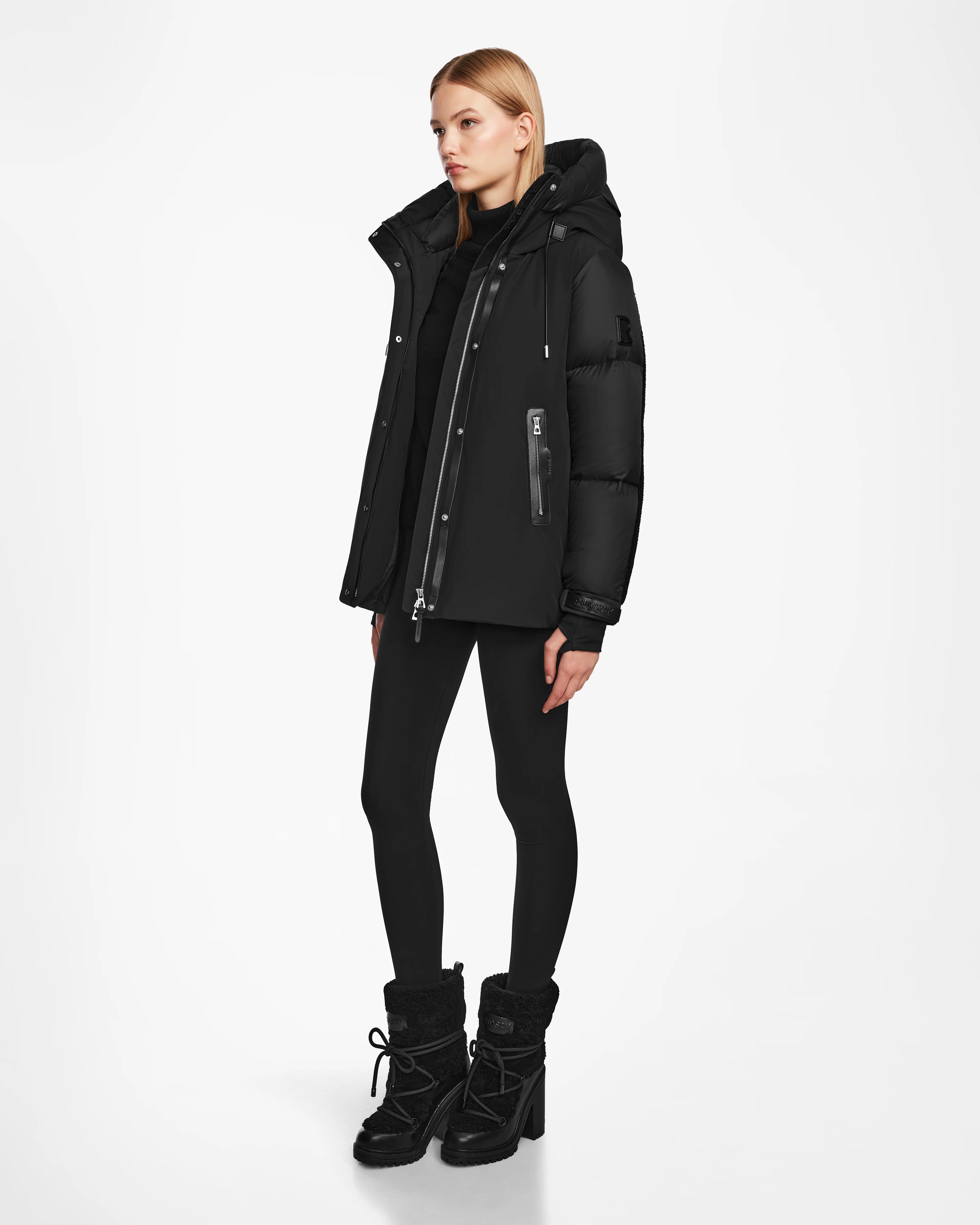 A woman with straight blonde hair stands against a white background wearing the Rudsak LOLI - 8124507 Black winter parka, paired with black leggings and boots. She looks slightly to the side with a neutral expression.