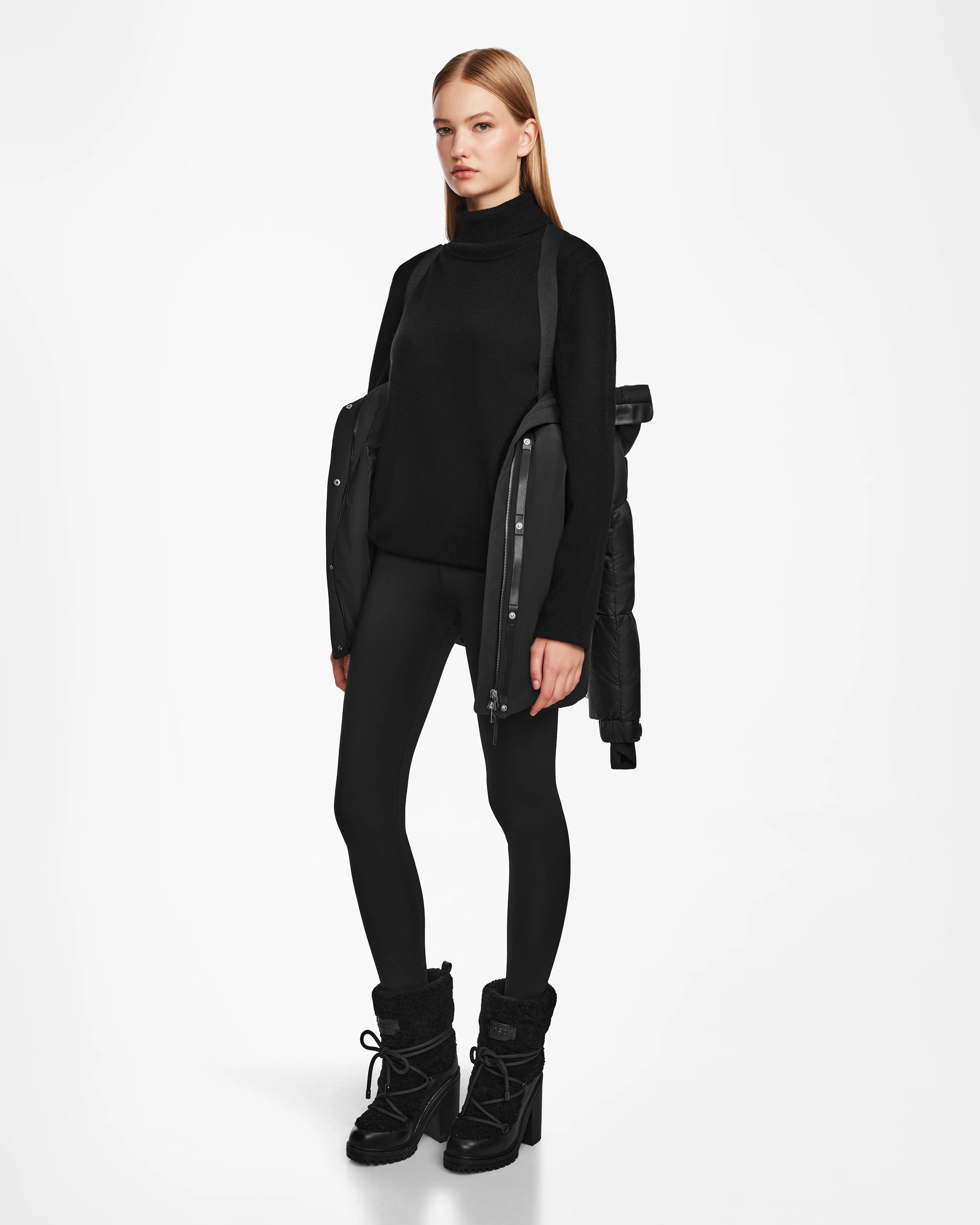 A woman stands against a white background wearing the Rudsak LOLI - 8124507 Black womens down parka draped over her shoulders, paired with a black turtleneck, leggings, and winter boots. She appears calm and looks straight ahead.