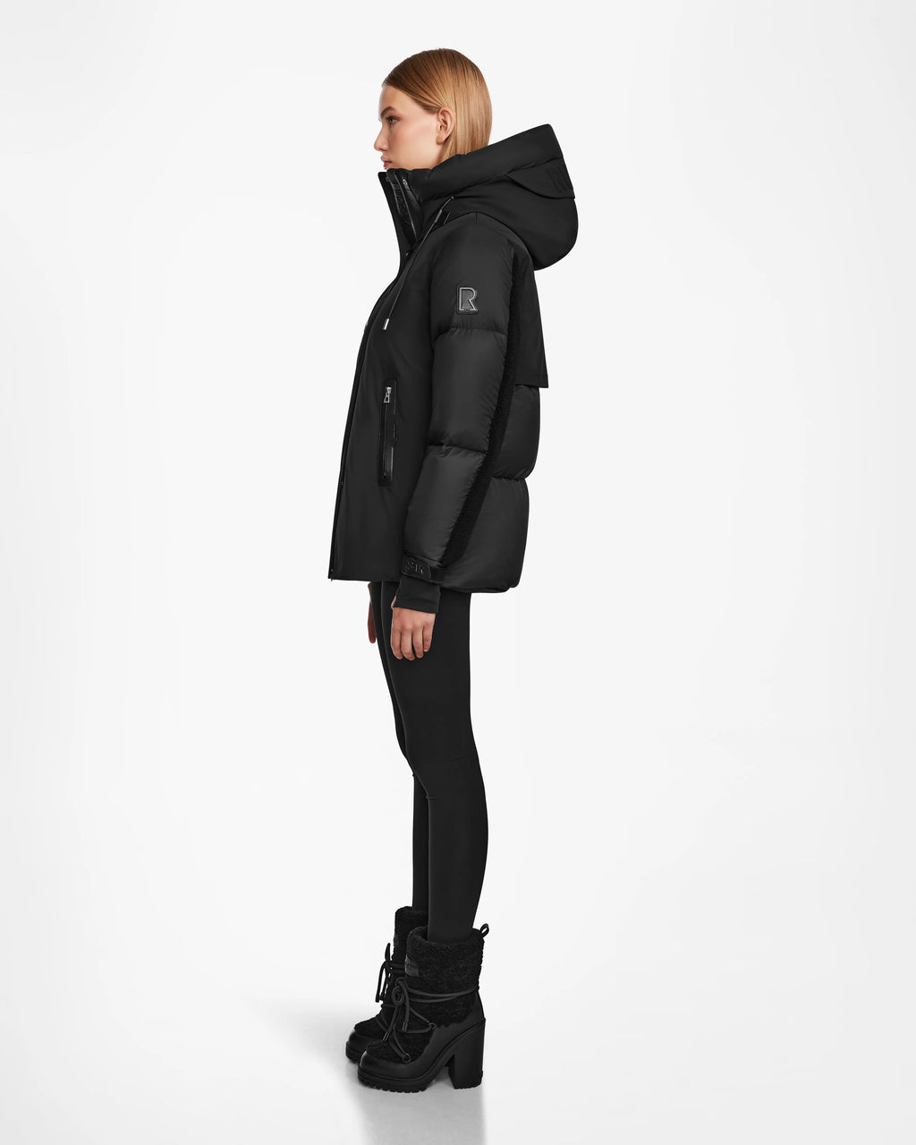 A woman stands in profile against a white background, wearing the LOLI - 8124507 Black womens down parka by Rudsak with a hood, paired with black leggings and black heeled winter boots.