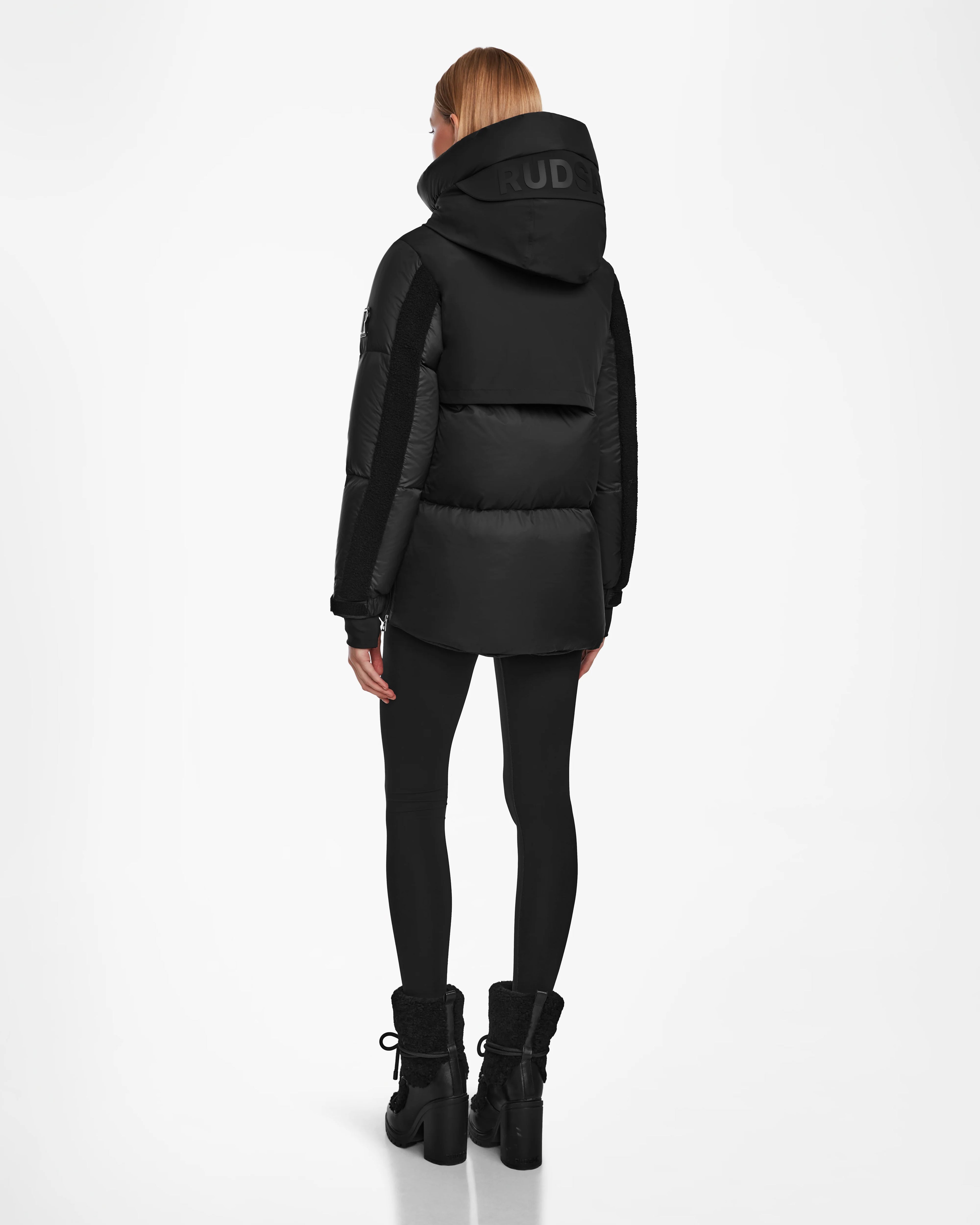 A woman with straight blonde hair is seen from behind wearing the Rudsak LOLI - 8124507 Black womens down parka, black leggings, and black lace-up boots against a plain white background.