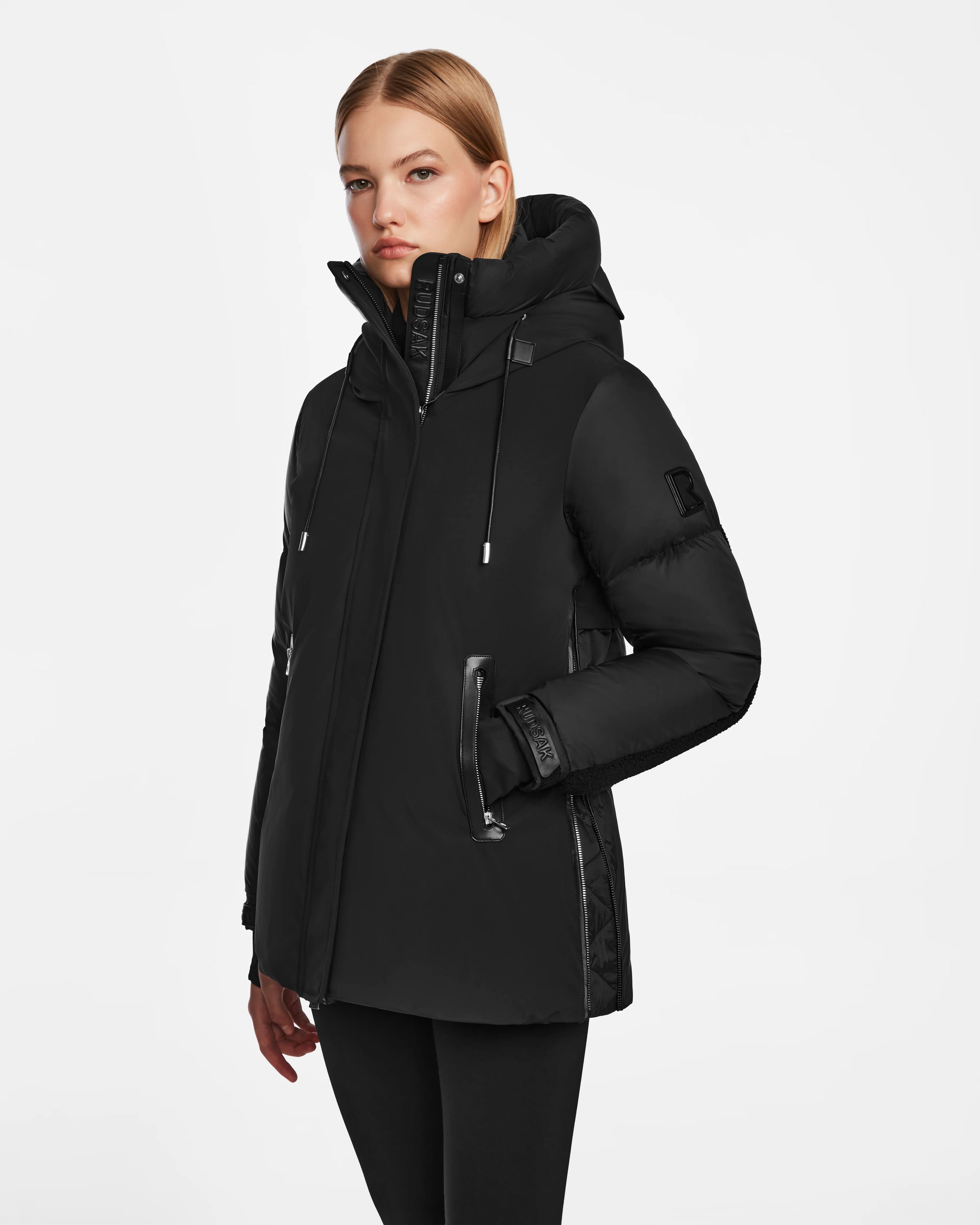 A person with straight blonde hair wears the Rudsak LOLI - 8124507 Black womens down parka, featuring zipper details, faux shearling accents, and side pockets, standing against a plain white background.