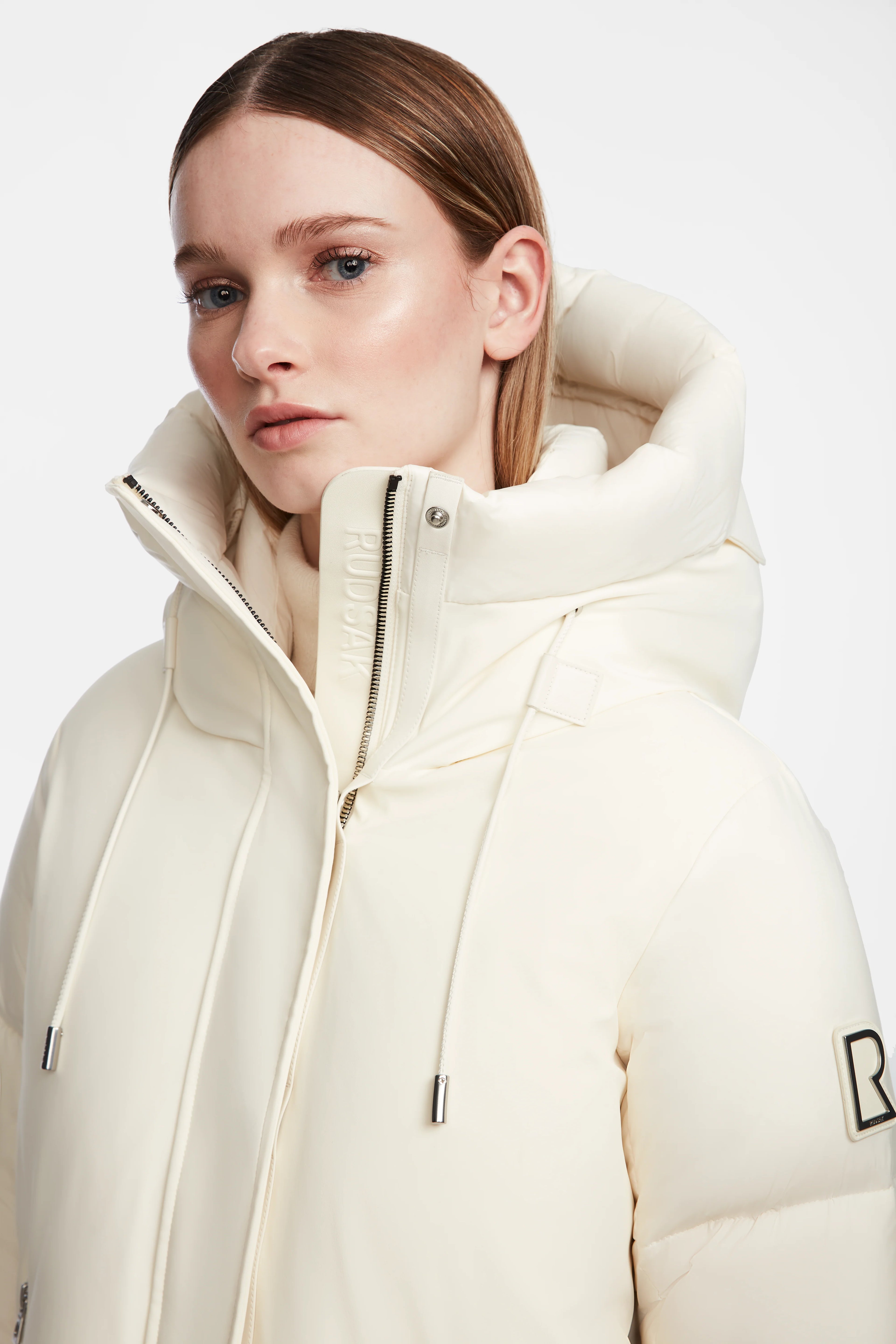 A woman wears the Rudsak LOLI - 8124507 Iceland off-white winter parka with a high collar, hood, drawstrings with metal tips, and a logo patch on the sleeve. She has straight light brown hair and a neutral expression.