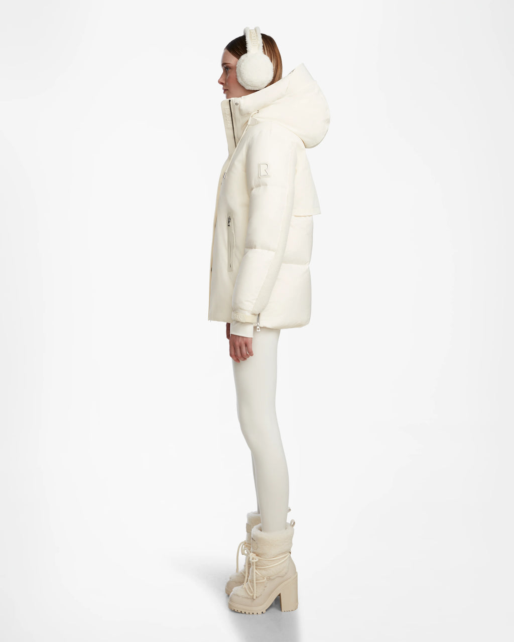 A woman in profile stands against a white background, wearing the Rudsak LOLI - 8124507 Iceland womens down parka with a hood, paired with white earmuffs, leggings, and fur-trimmed heeled winter boots.