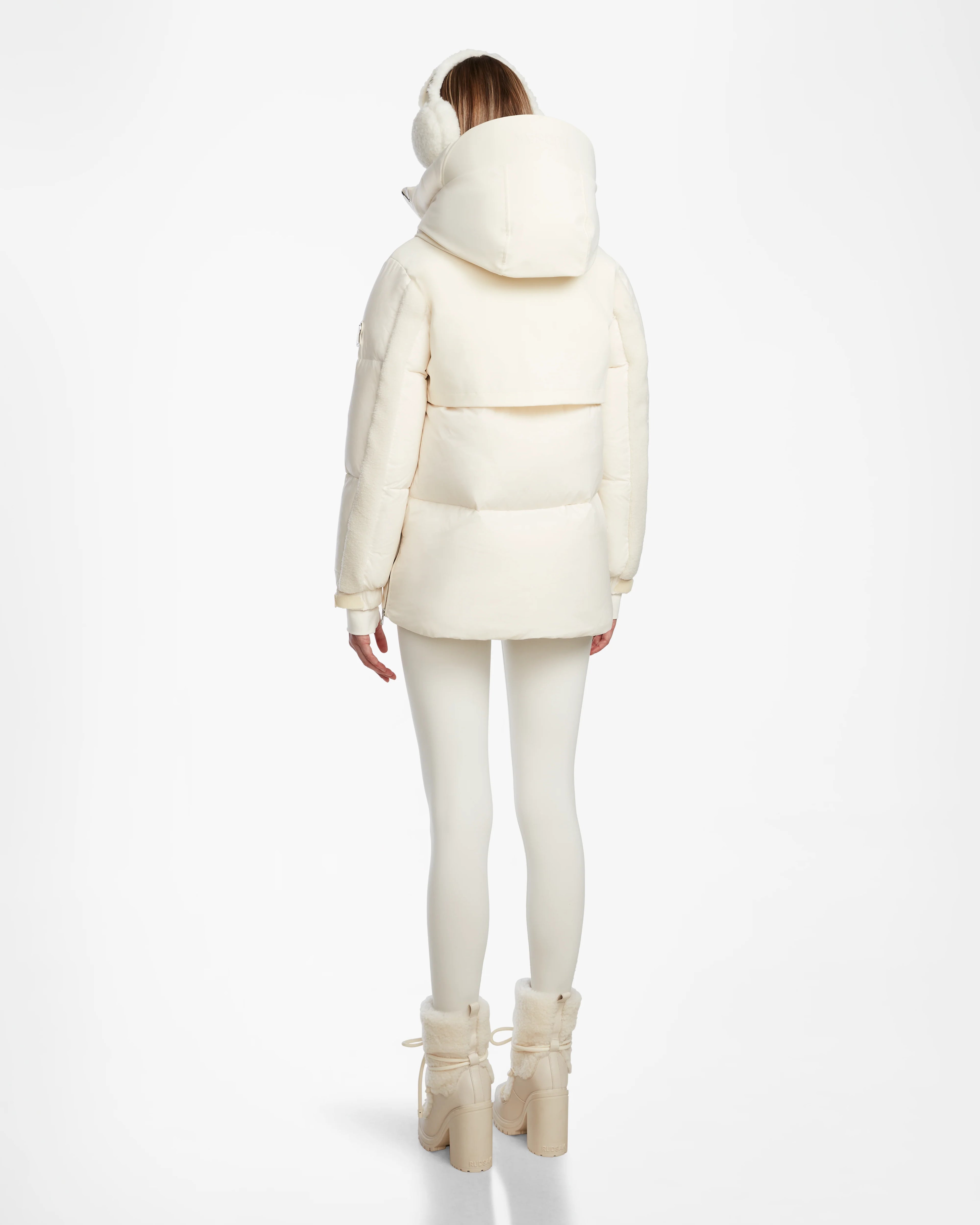 A person faces away from the camera wearing the cream-colored Rudsak LOLI - 8124507 Iceland womens down parka with a large hood, paired with matching off-white leggings, chunky boots, and earmuffs—all set against a white background.