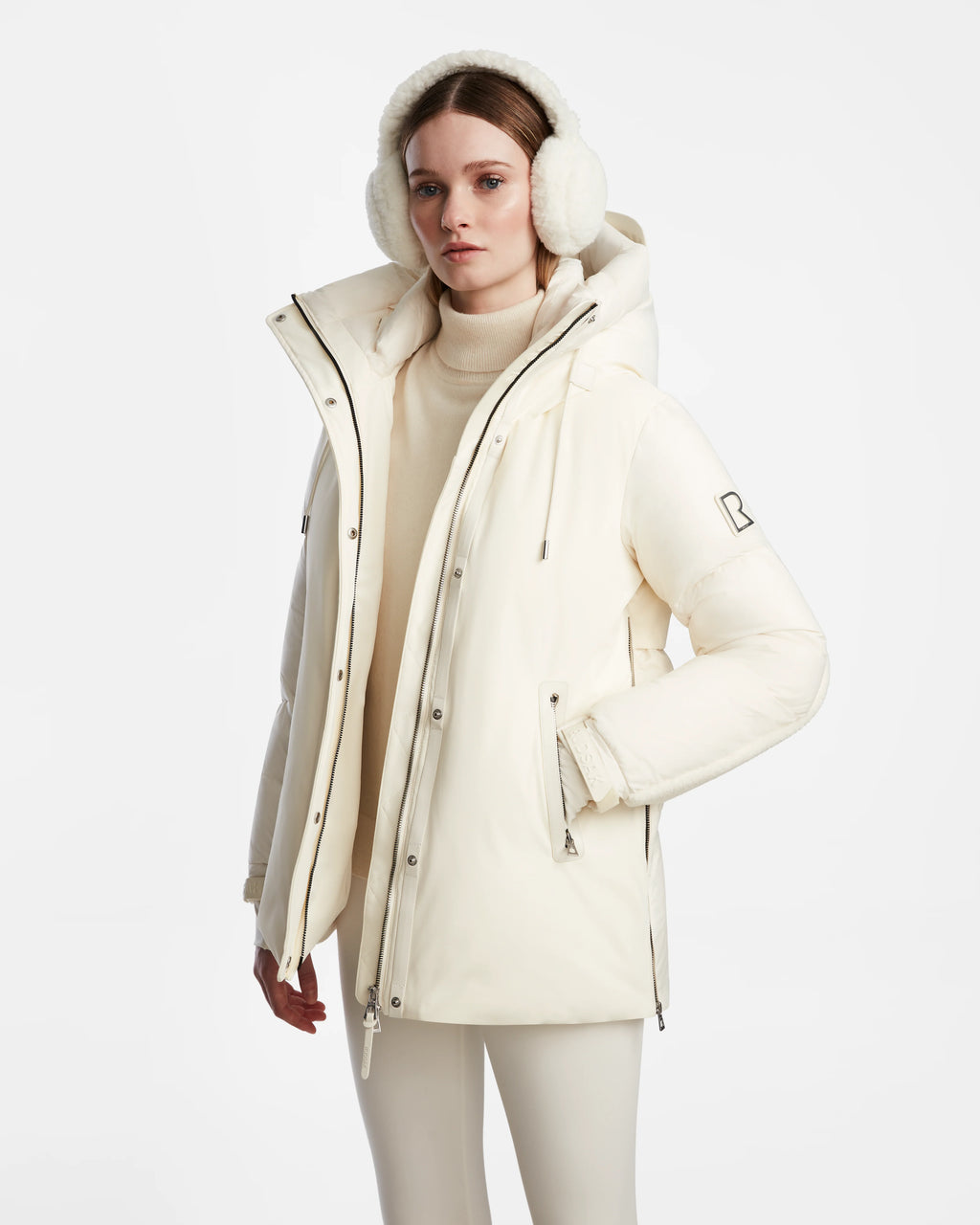 A woman stands against a white background wearing the Rudsak LOLI - 8124507 Iceland cream women’s down parka with a high collar, hood, faux shearling details, matching earmuffs, and light pants for warmth and style.