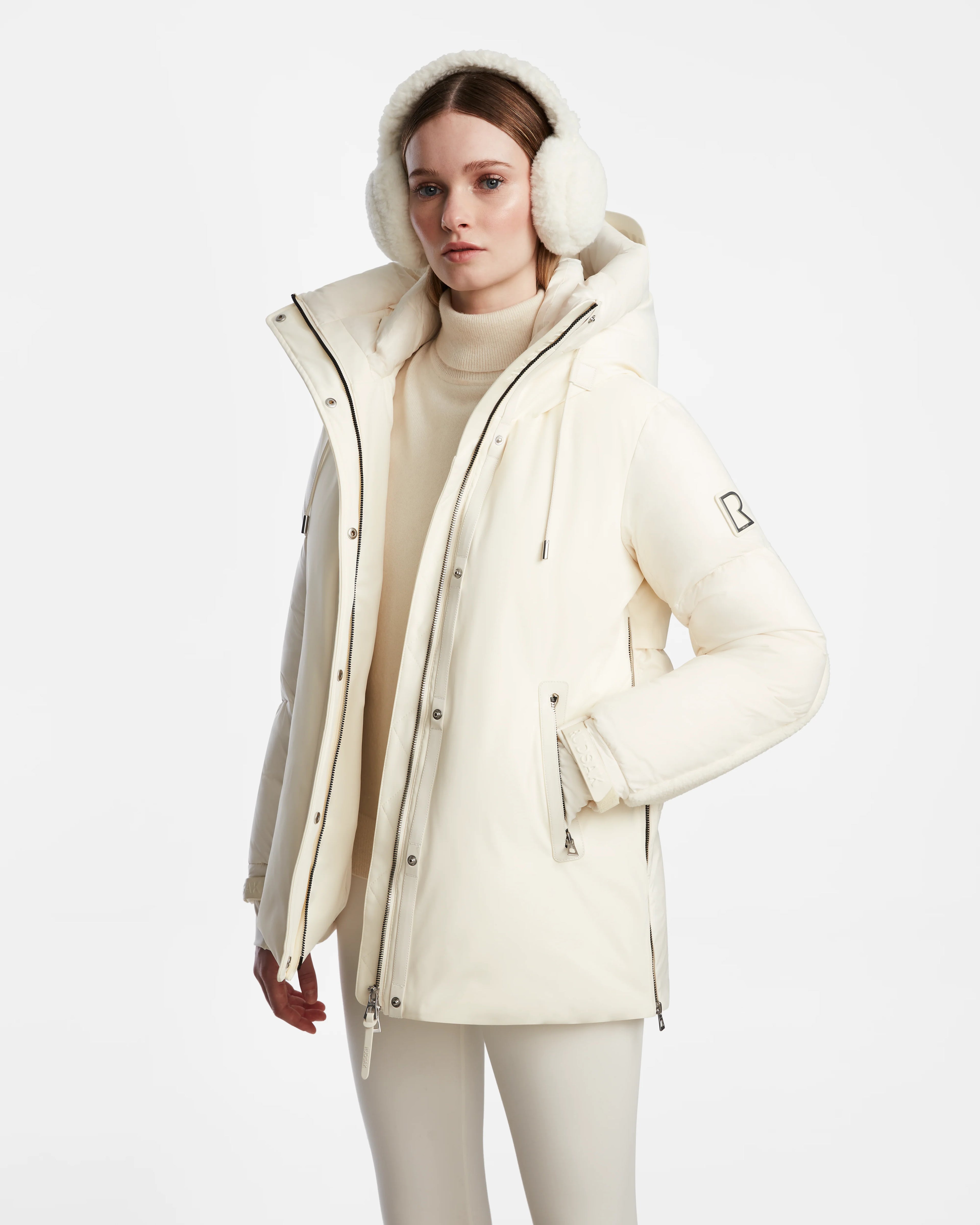 A woman stands against a white background wearing the Rudsak LOLI - 8124507 Iceland cream women’s down parka with a high collar, hood, faux shearling details, matching earmuffs, and light pants for warmth and style.