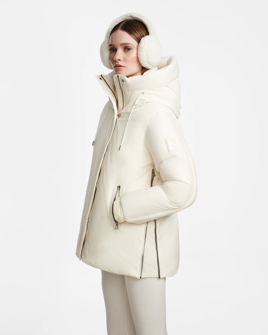 A woman in a Rudsak LOLI - 8124507 Iceland womens down parka with a high collar and large hood stands in profile, wearing matching white earmuffs and light pants against a plain white background.