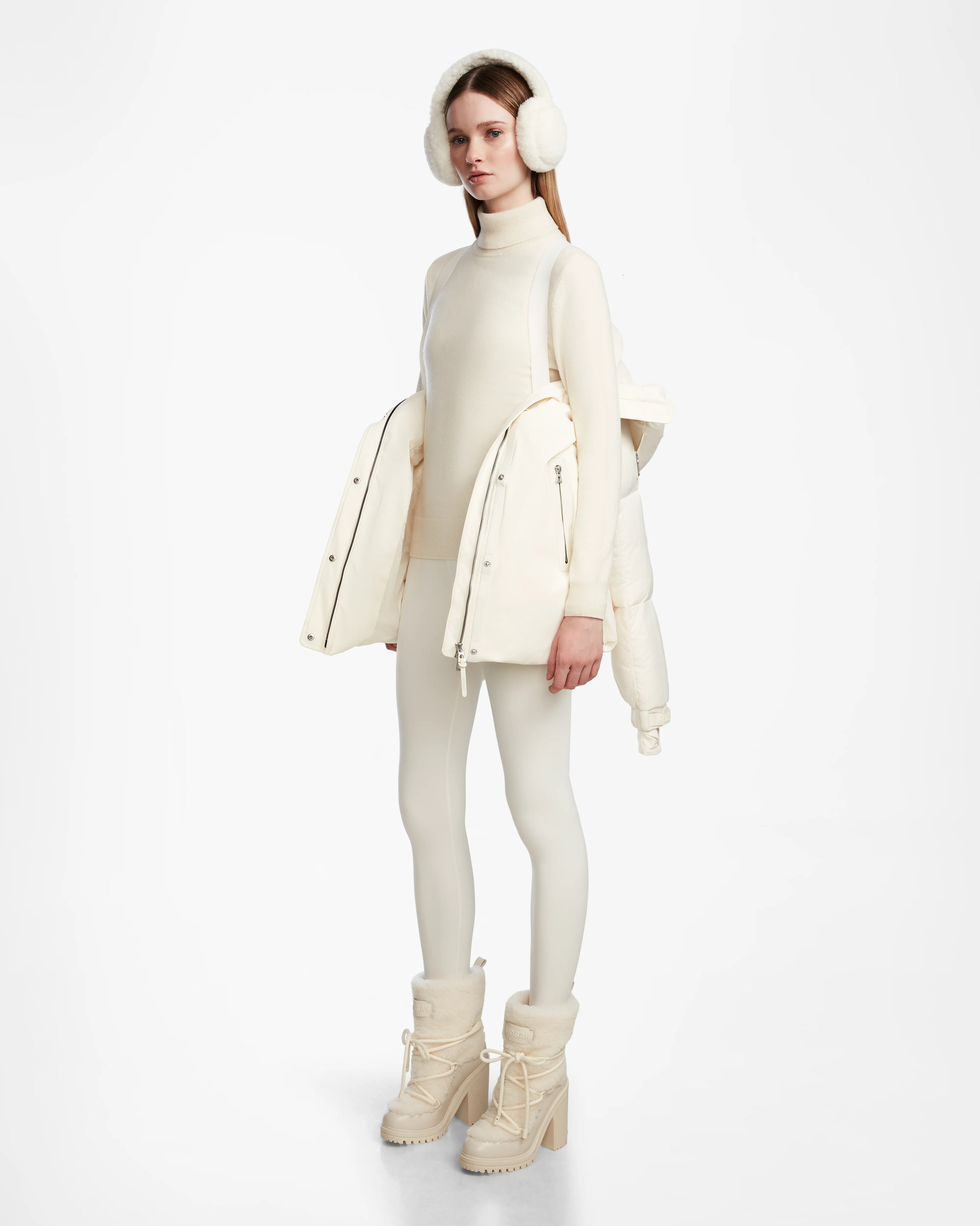A woman stands against a plain white background wearing an all-white winter outfit with earmuffs, a long-sleeve top, short skirt, opaque tights, lace-up boots, and the Rudsak LOLI - 8124507 Iceland down parka draped over her shoulders.