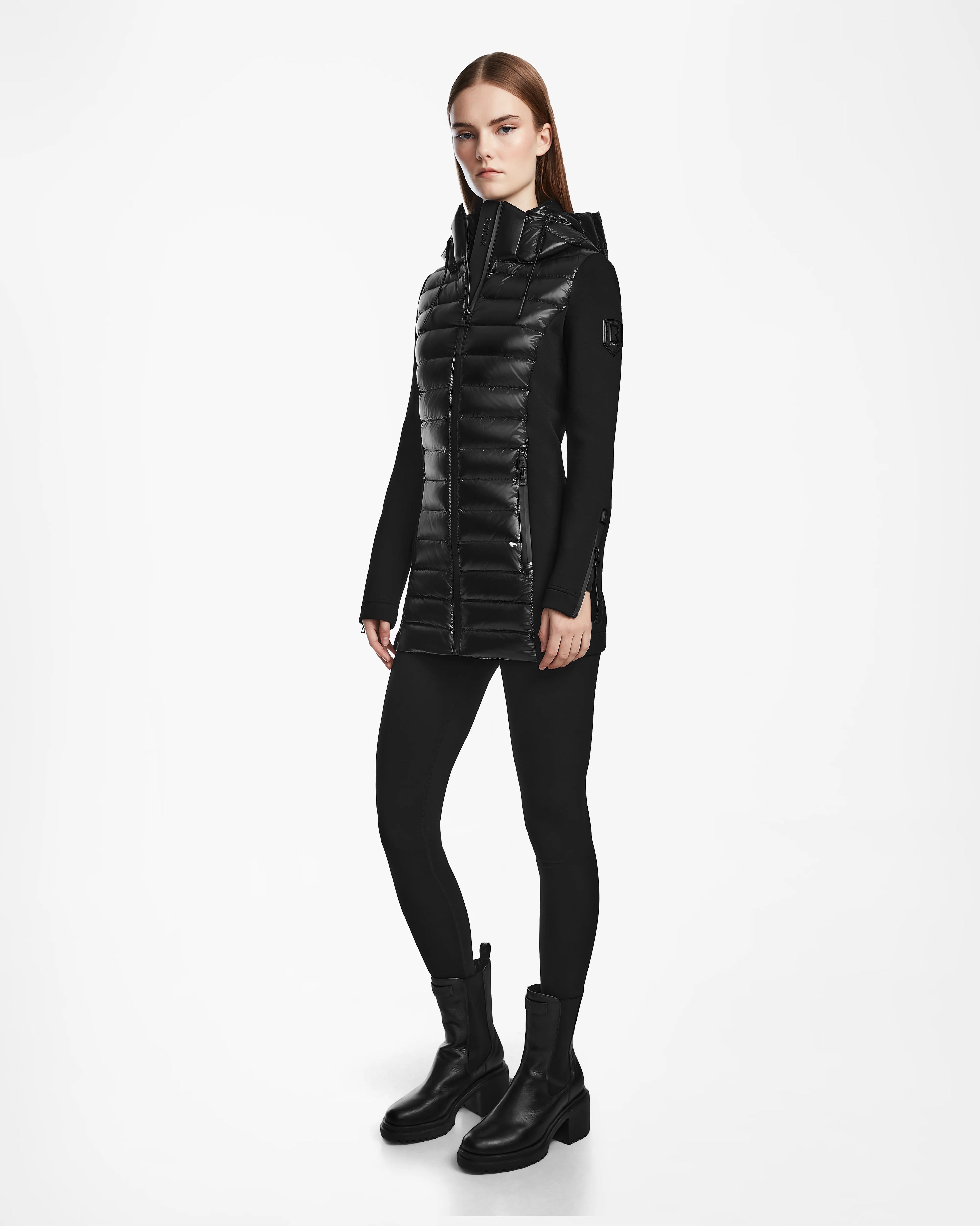 A woman with straight brown hair and a neutral expression stands against a white background wearing the Rudsak MARRY - L121003 BLACK puffer jacket over a black top, black leggings, and boots.