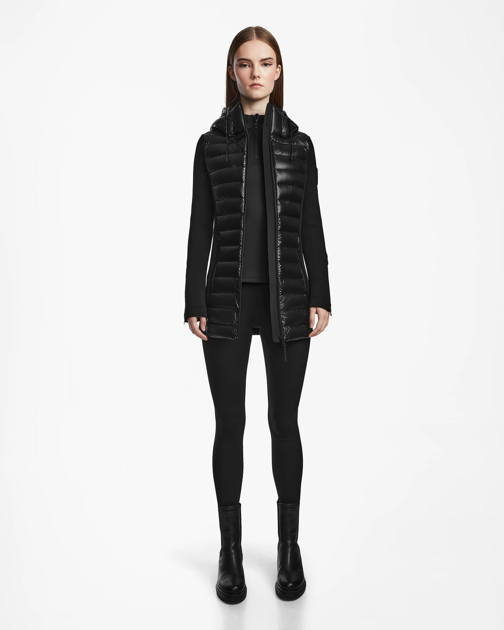 A woman faces forward against a white background, wearing the Rudsak MARRY - L121003 BLACK recycled down puffer vest over a black zip-up top, black leggings, and black ankle boots.
