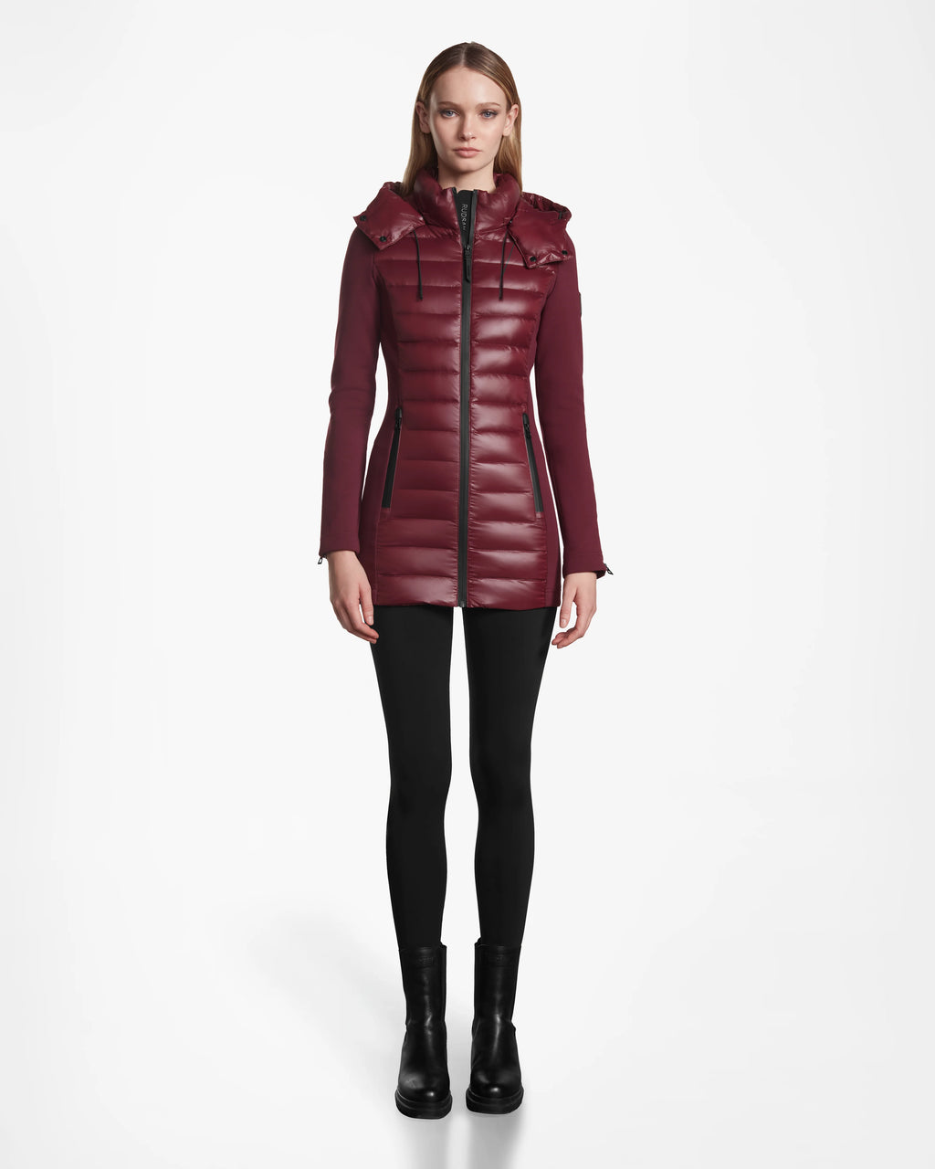 A woman faces forward, wearing the Rudsak MARRY - L121003 Wine long puffer with hood, black leggings, and black ankle boots against a plain white background.