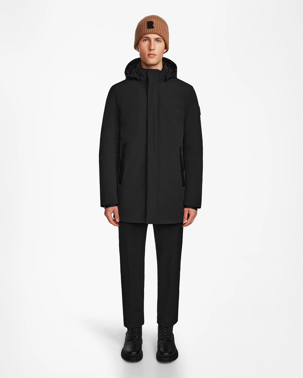 A person faces forward against a plain white background, wearing the Rudsak NATAN - M124519 Black mens parka, paired with black pants, black boots, and a brown knit beanie.