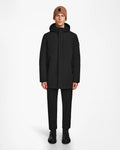 A person faces forward against a plain white background, wearing the Rudsak NATAN - M124519 Black mens parka, paired with black pants, black boots, and a brown knit beanie.