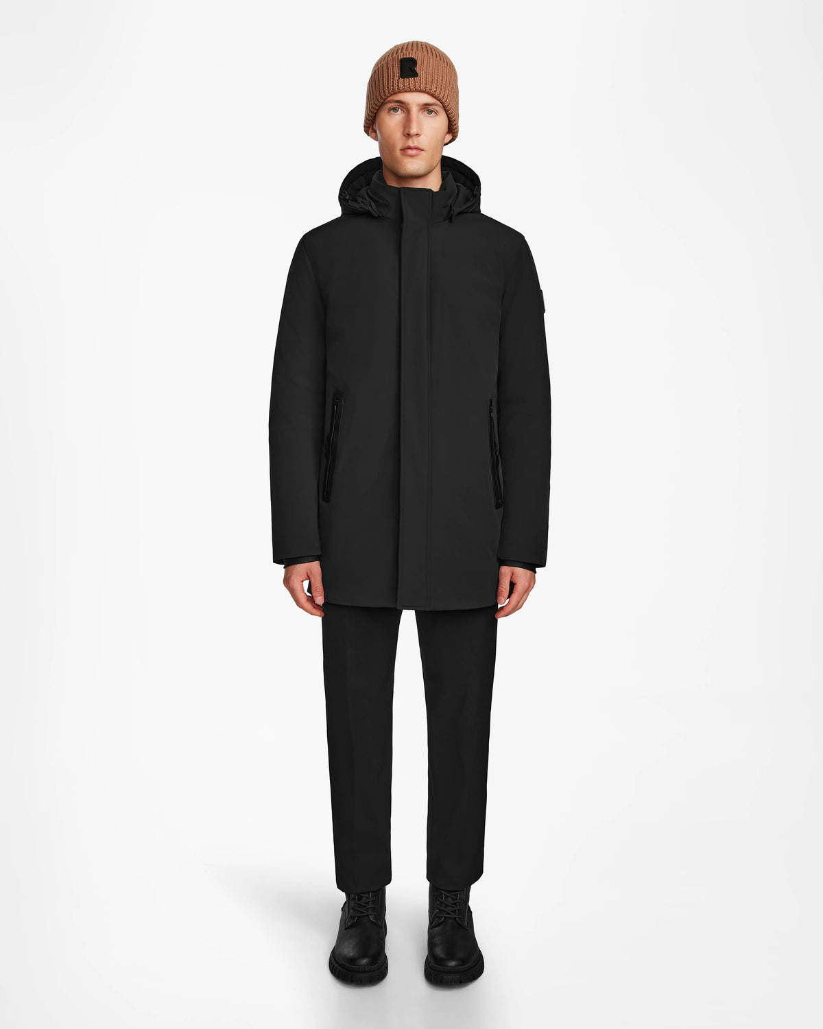 A person faces forward against a plain white background, wearing the Rudsak NATAN - M124519 Black mens parka, paired with black pants, black boots, and a brown knit beanie.