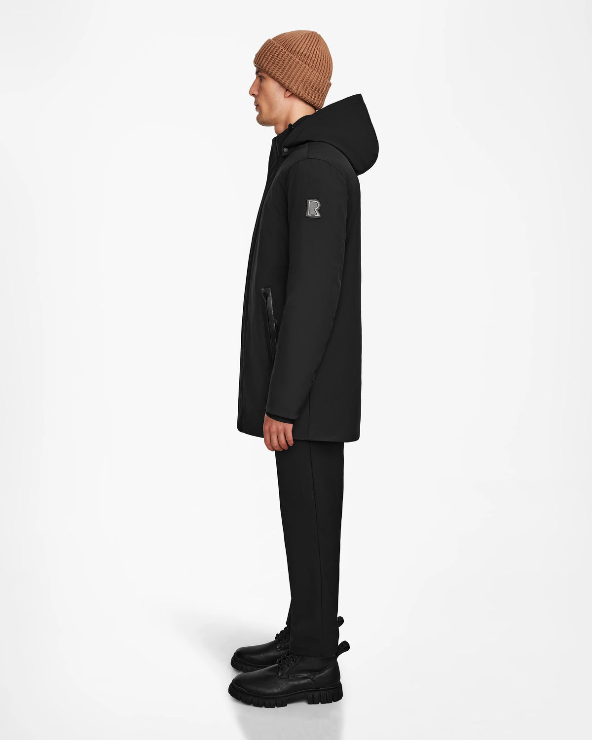 A person wearing a brown beanie, black pants, and black boots stands in profile against a white background, showcasing the Rudsak NATAN - M124519 Black men’s parka with down fill insulation.