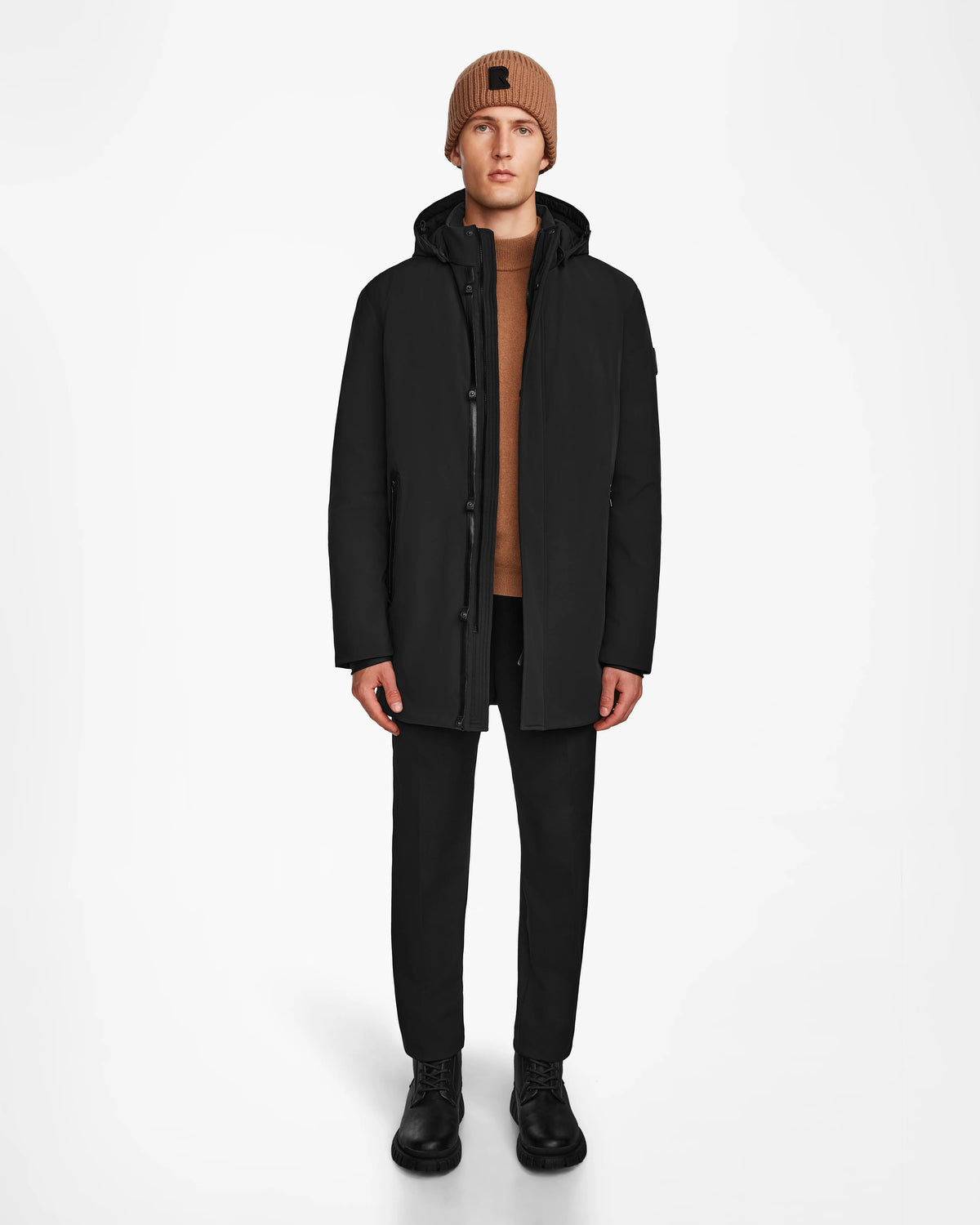 A person faces forward against a plain white background, wearing a brown knit beanie, brown sweater, black pants and shoes, and the Rudsak NATAN - M124519 Black men’s down-filled parka.
