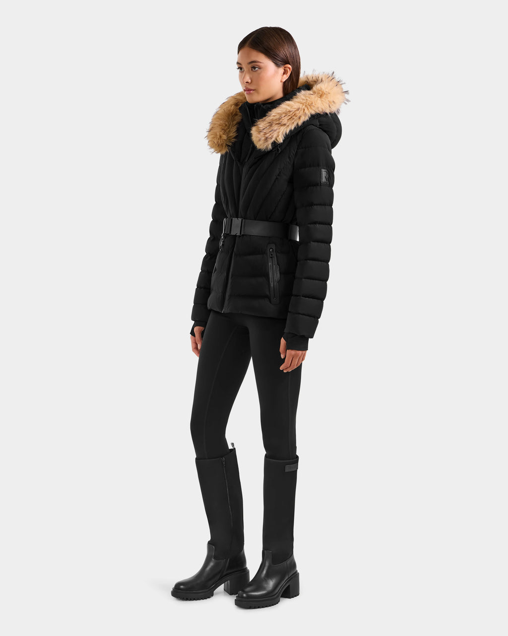 A woman wears the Rudsak ALISA F - L125527 Black quilted puffer coat with belted waist and faux-fur trim hood, styled with black leggings and knee-high boots against a plain background.
