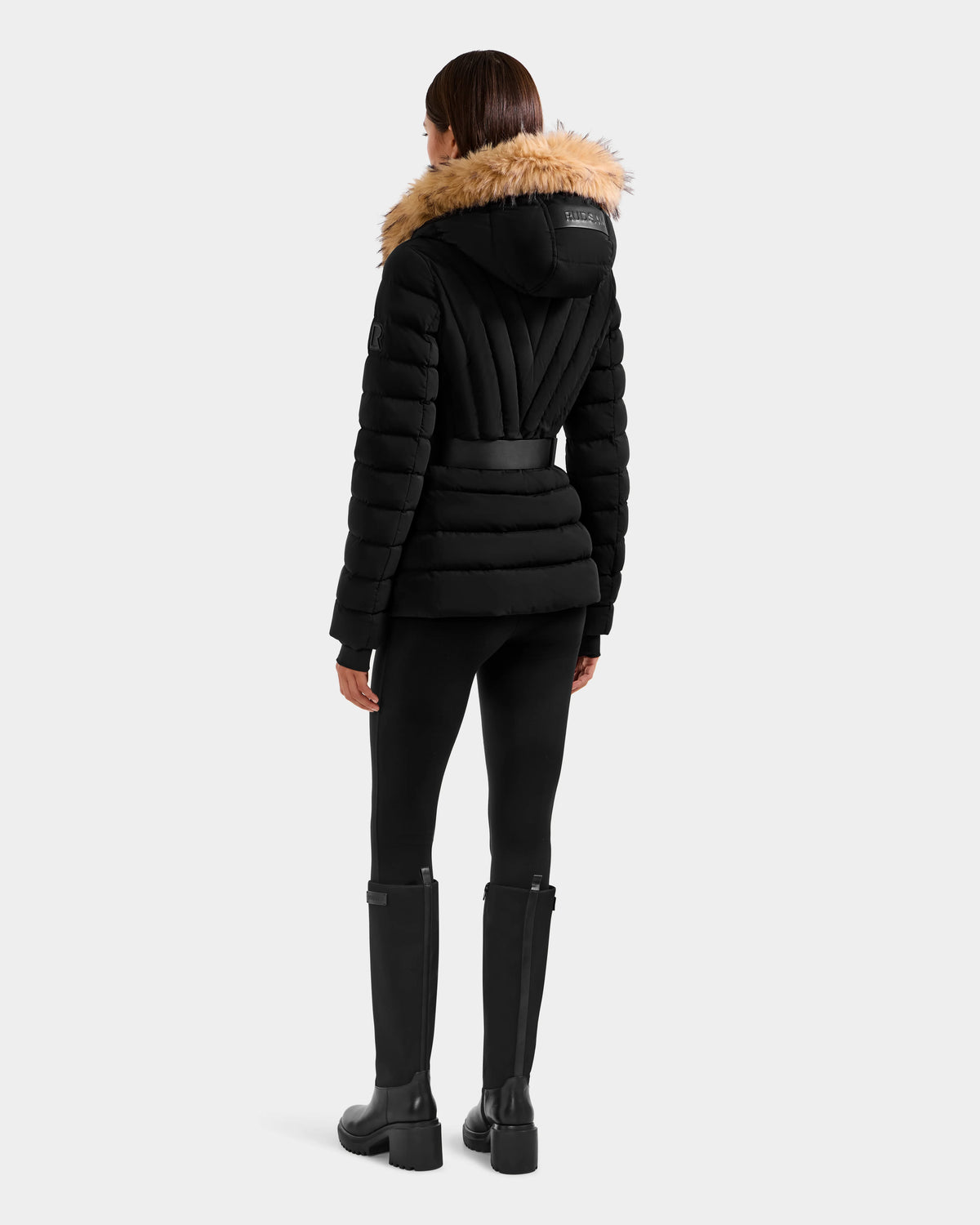 A woman faces away, wearing the Rudsak ALISA F - L125527 Black quilted puffer jacket with a faux-fur trim hood, paired with black leggings and tall black boots. The background is plain light gray.