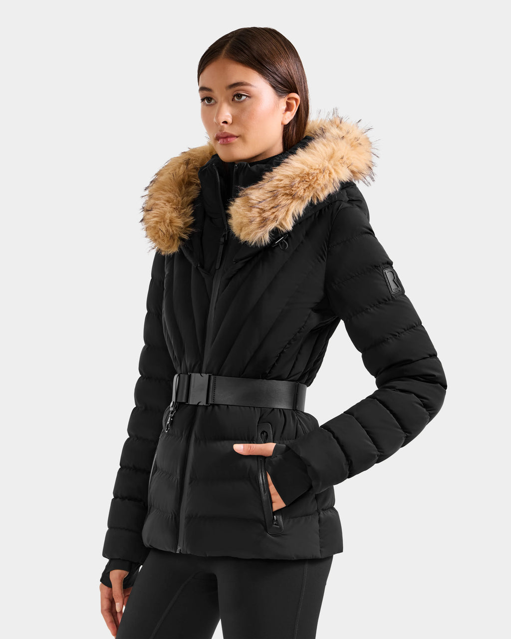 A woman with straight brown hair wears the Rudsak ALISA F - L125527 Black quilted puffer jacket, featuring a vegan-leather belt and a faux fur-trimmed hood, standing against a light gray background and looking slightly left.