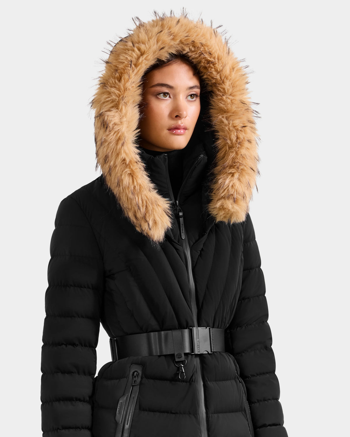 A woman wears the Rudsak ALISA F - L125527 Black belted quilted puffer coat with a large hood and tan faux-fur trim, standing against a plain light background.