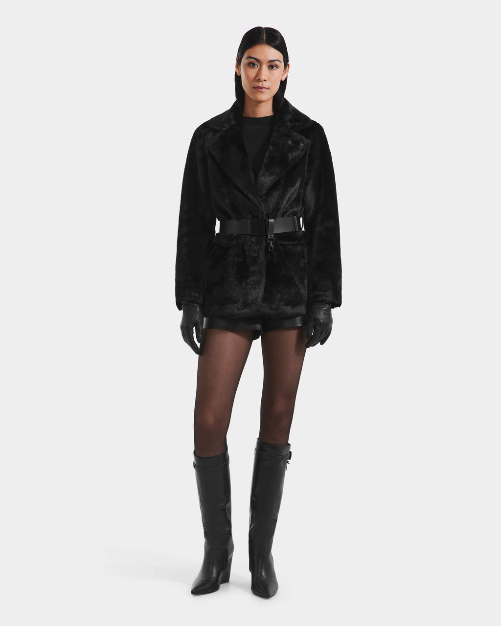 A woman stands facing forward in the Rudsak ASPEN F - 8125604 Black women’s faux fur belted jacket, styled with black shorts, sheer tights, knee-high boots, and gloves against a plain white background.
