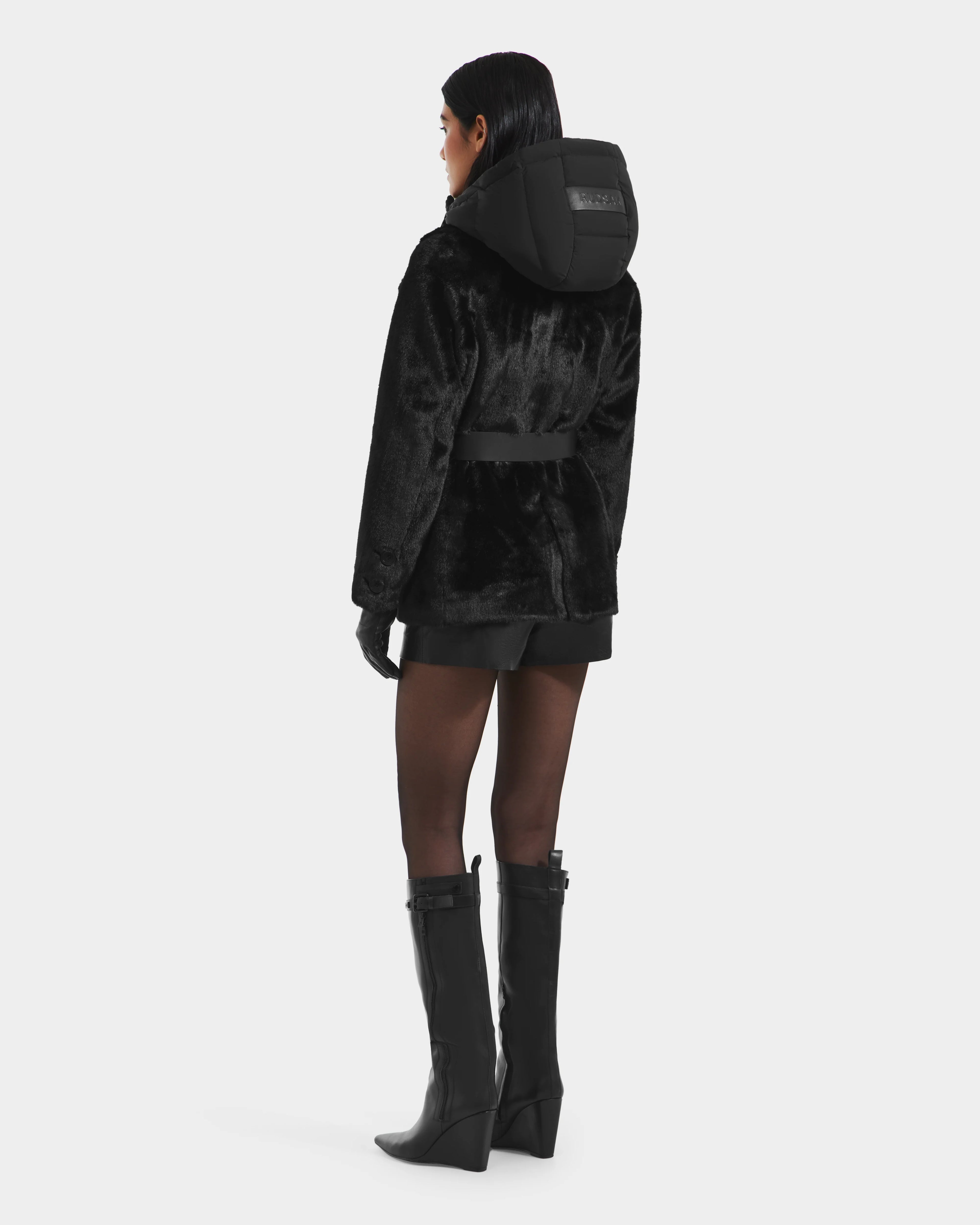 A woman faces away wearing the Rudsak ASPEN F - 8125604 Black faux fur jacket with a belted waist and hood, paired with black gloves, shorts, and knee-high boots—a chic, cold-weather look. The jacket sports a branded label on the hood.