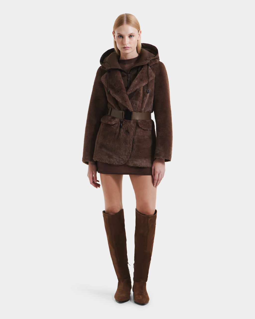 A woman faces forward in the Rudsak ASPEN F - 8125604 Mocha, a brown belted faux fur jacket with a removable down-insulated hood. She pairs it with a short brown dress and knee-high suede boots against a white background.