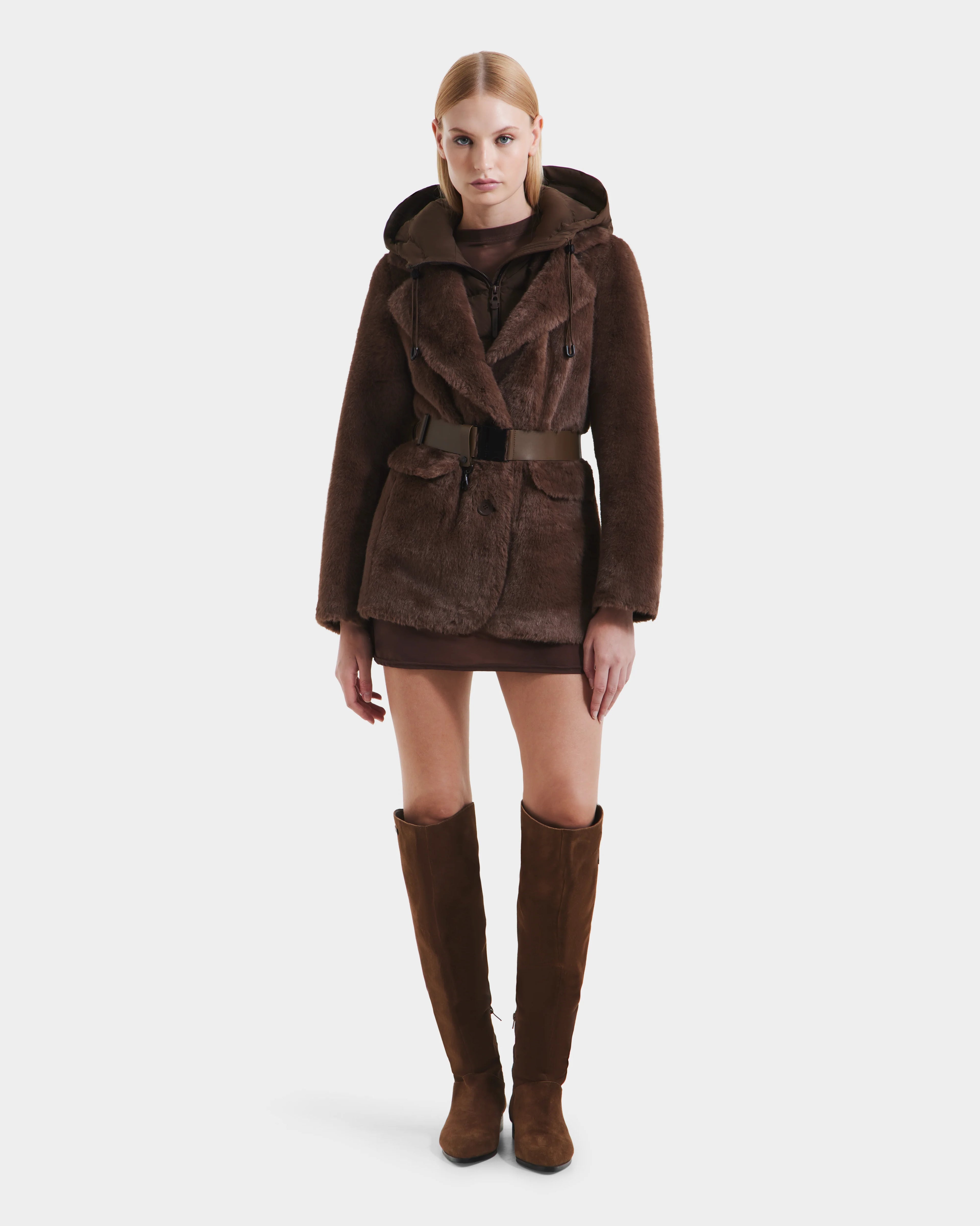 A woman faces forward in the Rudsak ASPEN F - 8125604 Mocha, a brown belted faux fur jacket with a removable down-insulated hood. She pairs it with a short brown dress and knee-high suede boots against a white background.