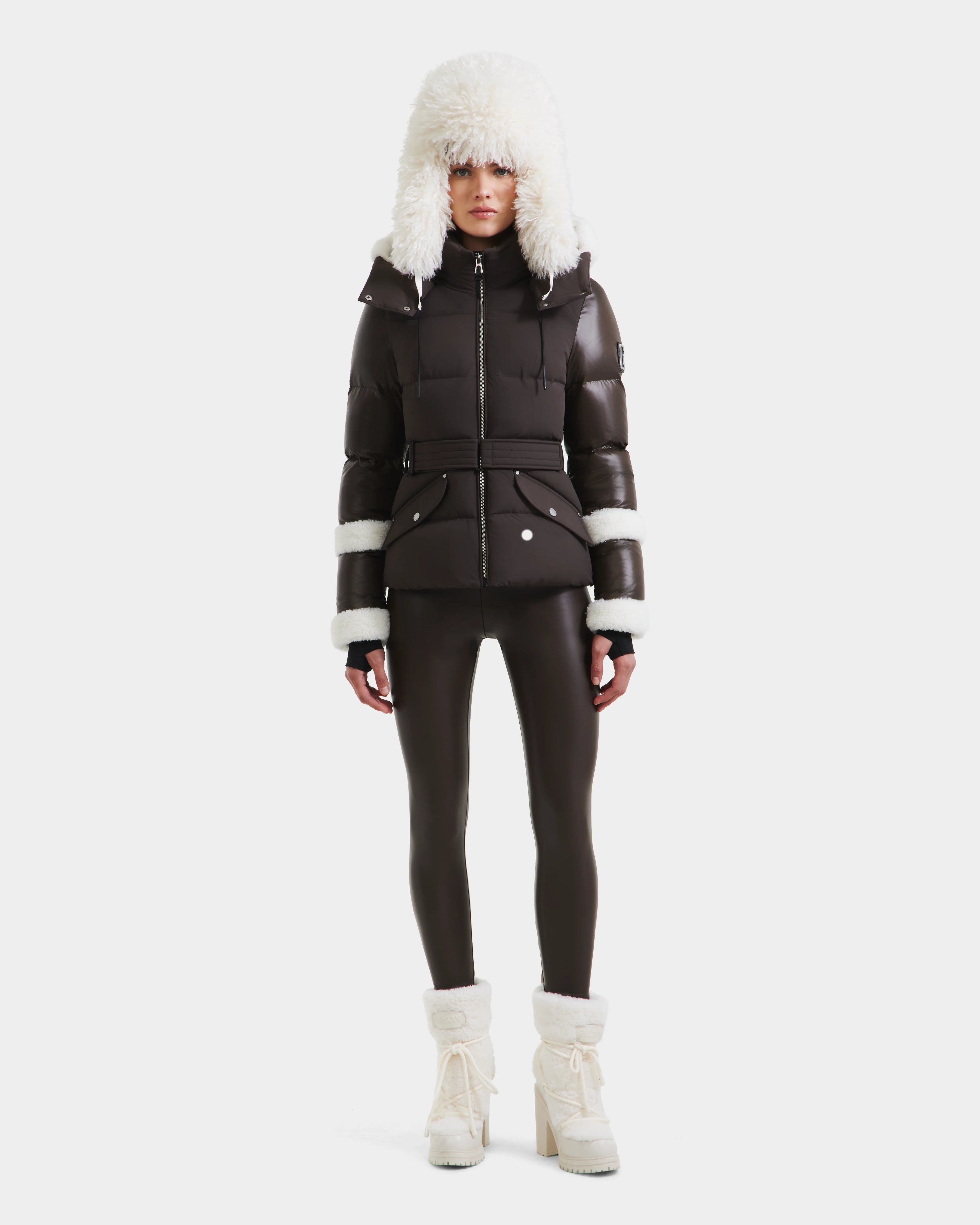 A person faces forward against a white background, wearing the Rudsak AUDREY - 8124582 Espresso puffer jacket with recycled Stormshell fabric and white fur trim, paired with matching dark leggings and white fur-lined boots.