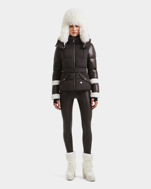 A person faces forward against a white background, wearing the Rudsak AUDREY - 8124582 Espresso puffer jacket with recycled Stormshell fabric and white fur trim, paired with matching dark leggings and white fur-lined boots.