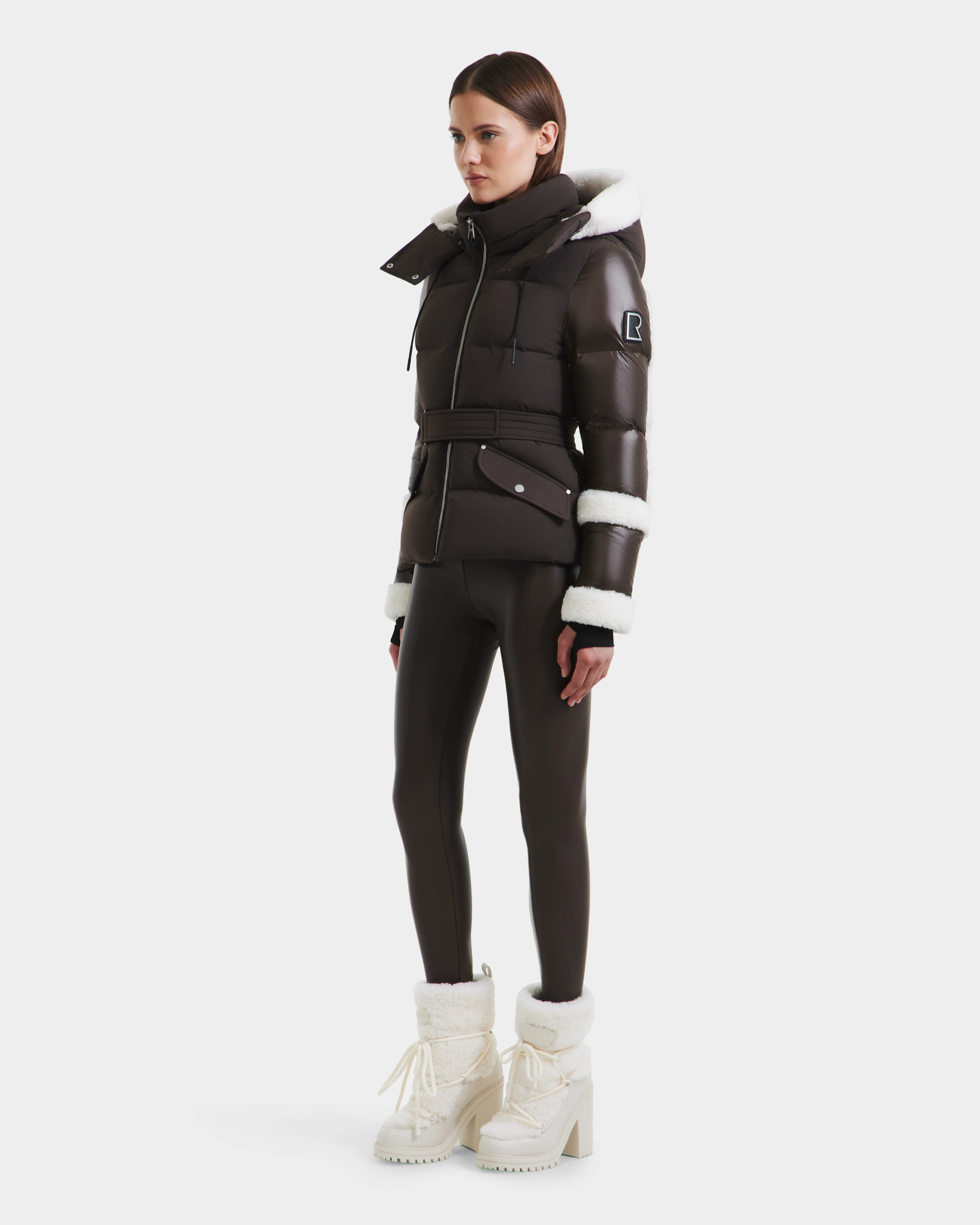 A woman models the Rudsak AUDREY - 8124582 Espresso brown down puffer jacket with white faux fur trim, paired with matching leggings and fur-lined boots against a plain white background.