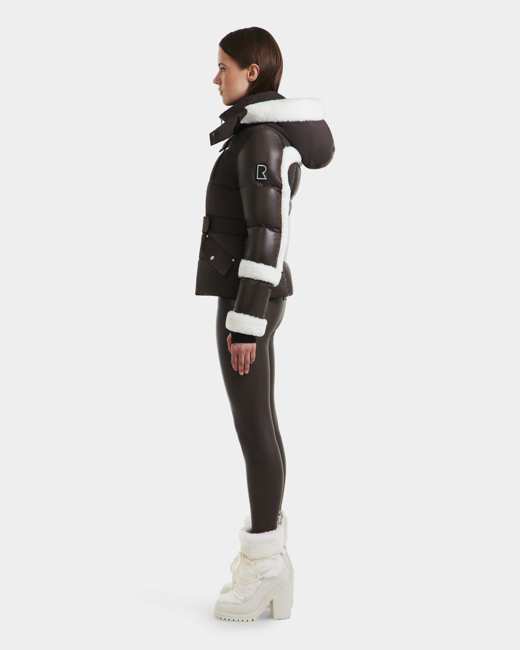 A woman stands in profile wearing the Rudsak AUDREY - 8124582 Espresso down puffer jacket, made from recycled Stormshell fabric with white fur trim and hood. She pairs it with black leggings and white platform boots; a patch details the sleeve.