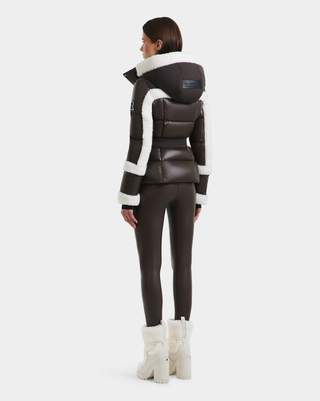 A woman faces away, wearing Rudsaks AUDREY - 8124582 Espresso dark brown down puffer with white shearling trim, crafted from recycled Stormshell fabric, paired with fitted pants and fur-lined boots against a plain white background.