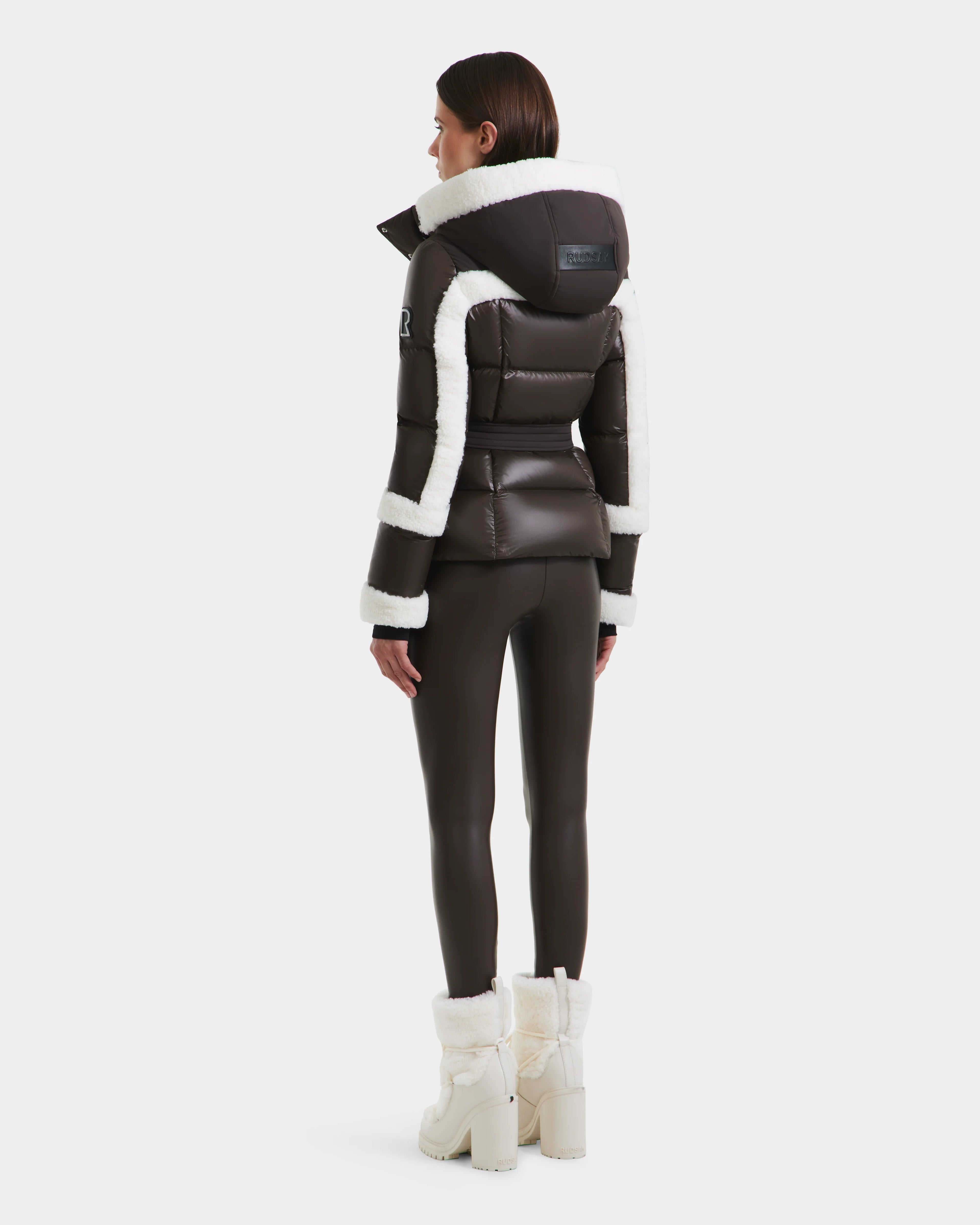 A woman faces away, wearing Rudsaks AUDREY - 8124582 Espresso dark brown down puffer with white shearling trim, crafted from recycled Stormshell fabric, paired with fitted pants and fur-lined boots against a plain white background.