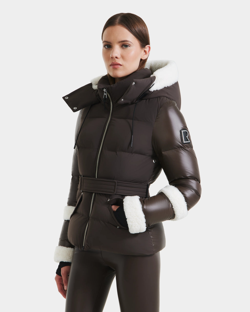 A woman stands against a white background wearing Rudsaks AUDREY - 8124582 Espresso, a dark brown belted down puffer jacket with white faux fur trim and RDS certified down, paired with matching brown leggings.