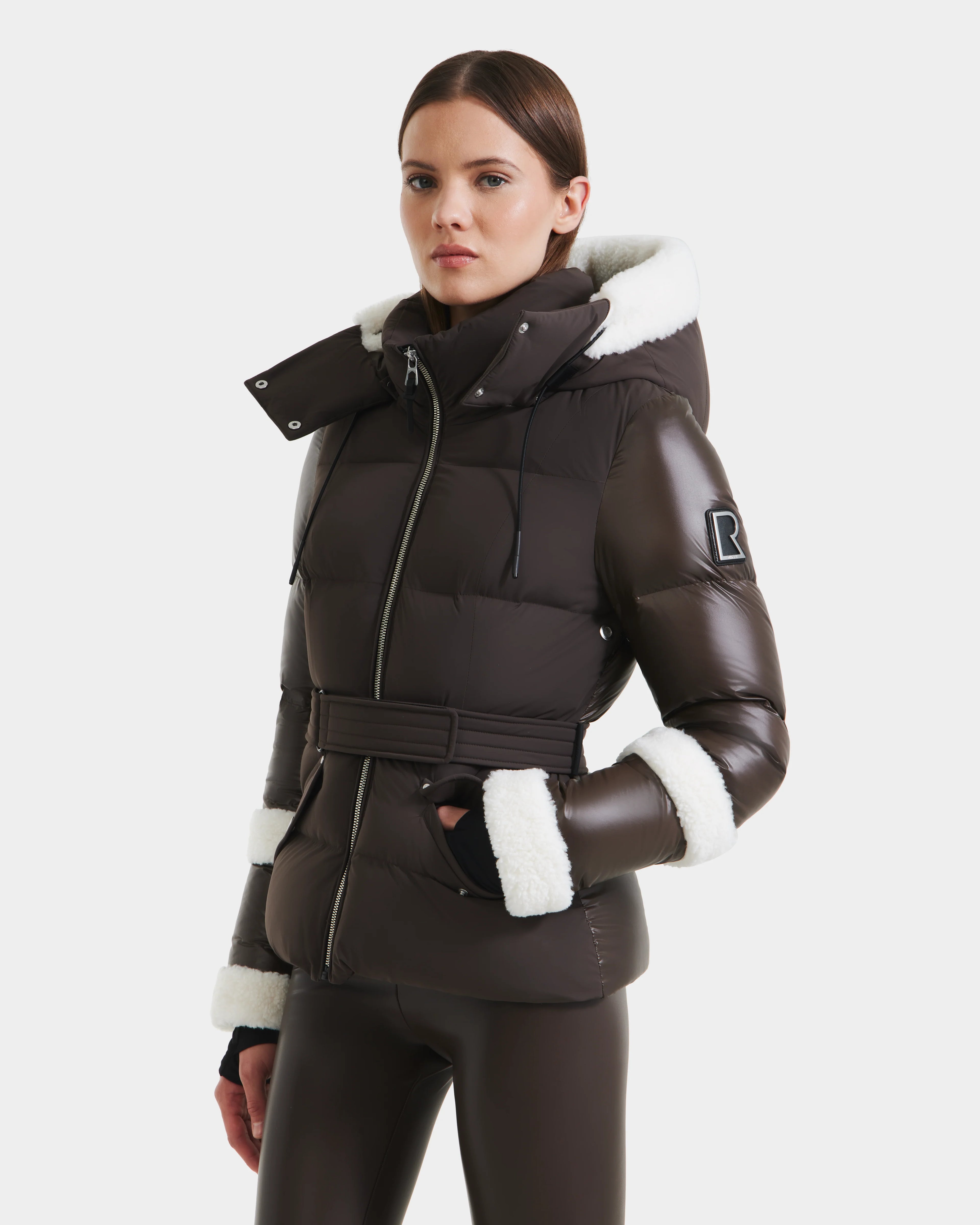 A woman stands against a white background wearing Rudsaks AUDREY - 8124582 Espresso, a dark brown belted down puffer jacket with white faux fur trim and RDS certified down, paired with matching brown leggings.