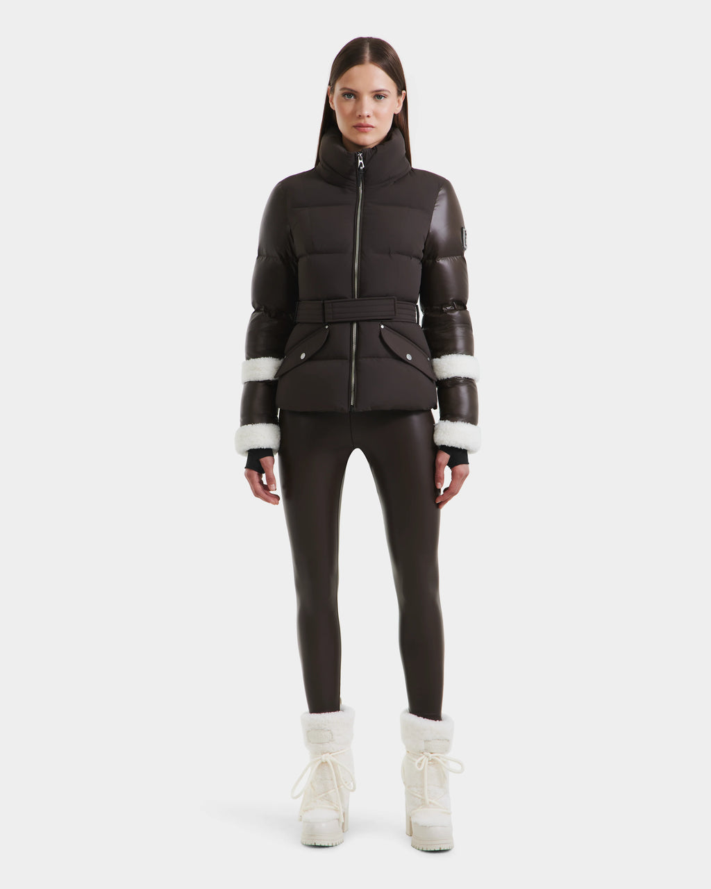 A woman stands facing forward in a Rudsak AUDREY - 8124582 Espresso down puffer jacket with white fur trim on the sleeves, black leggings, and white fur-lined winter boots against a plain white background.