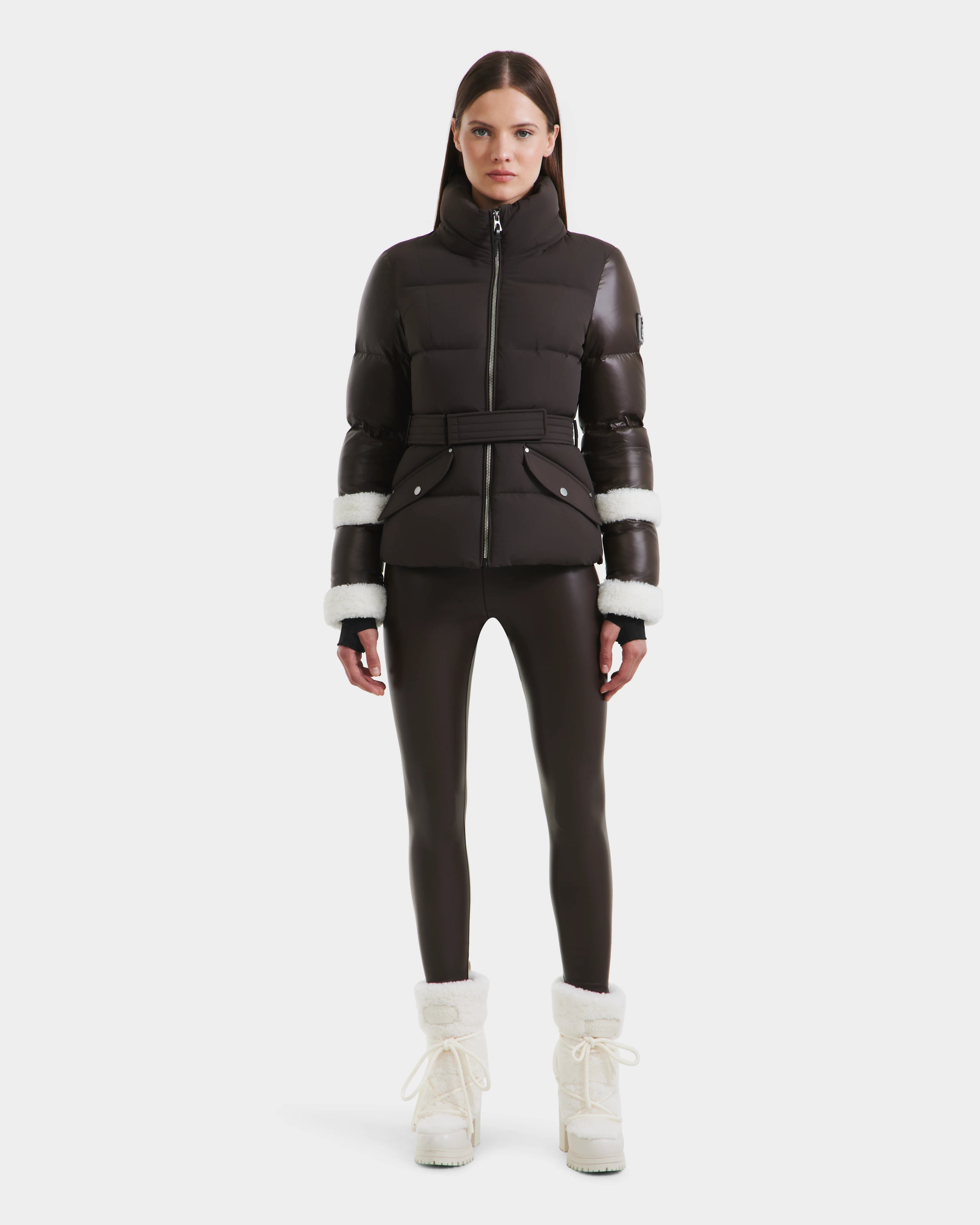 A woman stands facing forward in a Rudsak AUDREY - 8124582 Espresso down puffer jacket with white fur trim on the sleeves, black leggings, and white fur-lined winter boots against a plain white background.