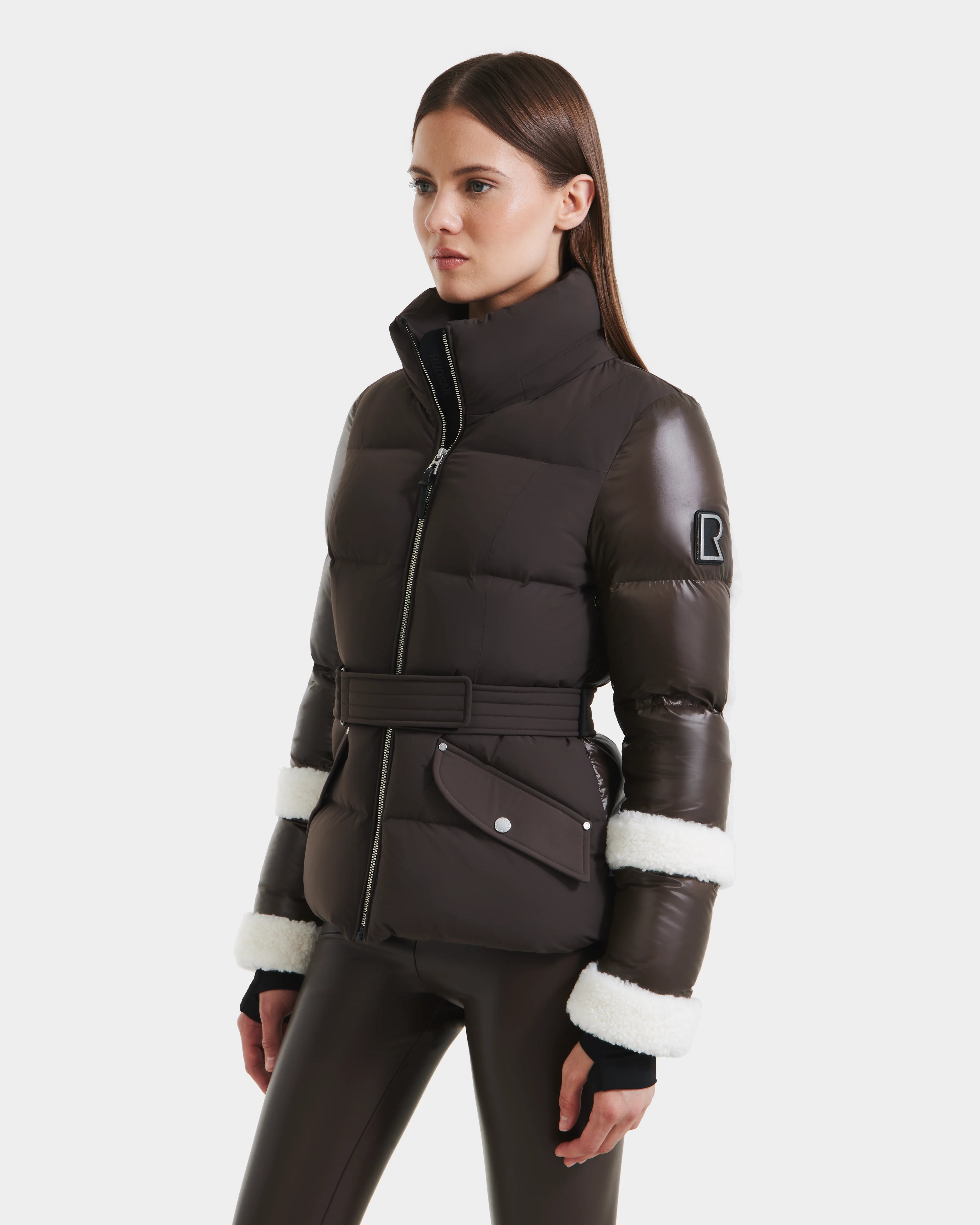 A woman models the Rudsak AUDREY - 8124582 Espresso, a dark brown puffer jacket with white fur sleeve trim, belted waist, and zippered pockets, paired with dark pants against a plain white background.