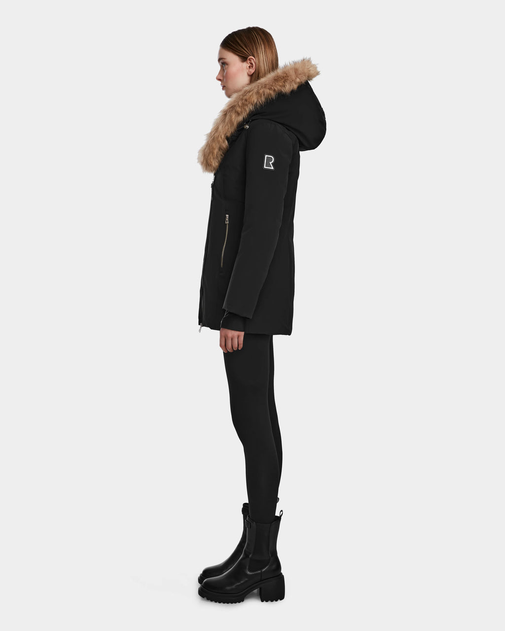 A woman stands in profile against a plain white background, wearing the Rudsak AURORE - L123561 Black/Natural fitted winter coat with a fur-trimmed hood, paired with black leggings and black boots.