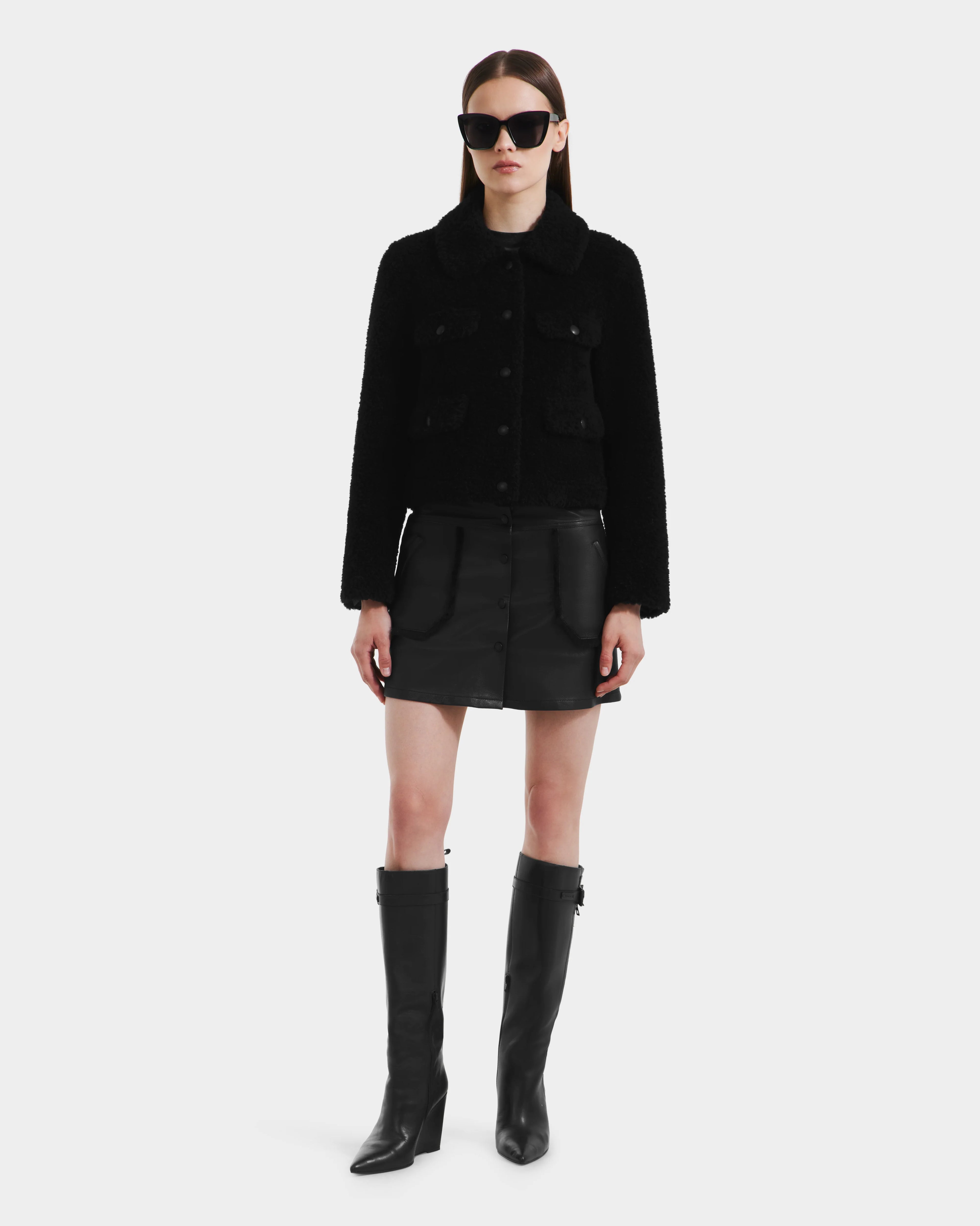 A woman faces forward against a white background, wearing the Rudsak BLAIR - 8125513 Black modern jacket, paired with a black mini skirt, tall black boots, and oversized black sunglasses.