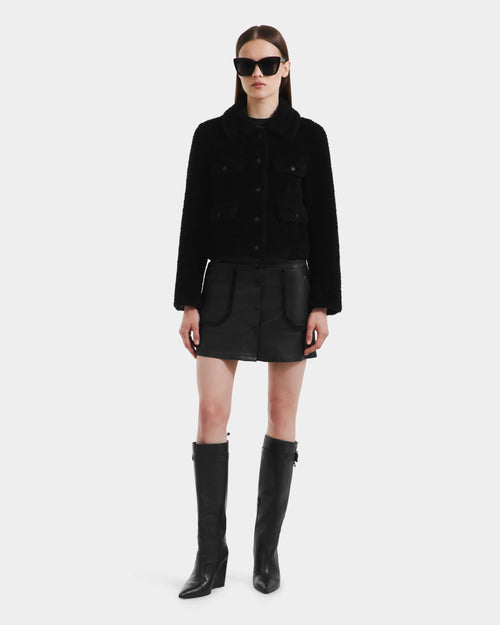 A woman faces forward against a white background, wearing the Rudsak BLAIR - 8125513 Black modern jacket, paired with a black mini skirt, tall black boots, and oversized black sunglasses.