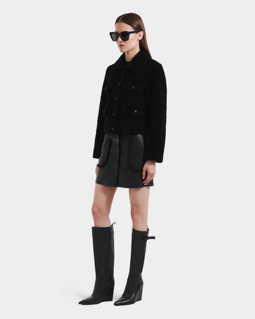 A woman wearing Rudsak’s BLAIR - 8125513 Black sunglasses, a cropped teddy jacket, black leather mini skirt, and black knee-high boots stands against a plain white background.