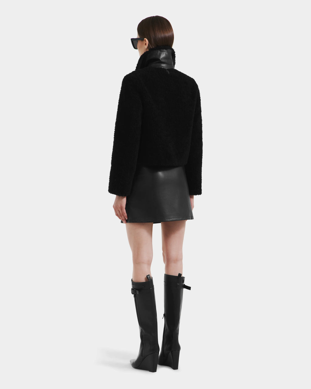 A woman with straight brown hair wears Rudsak’s BLAIR - 8125513 Black cropped teddy jacket, sunglasses, a black leather mini skirt, and black knee-high boots while facing away from the camera against a plain white background.