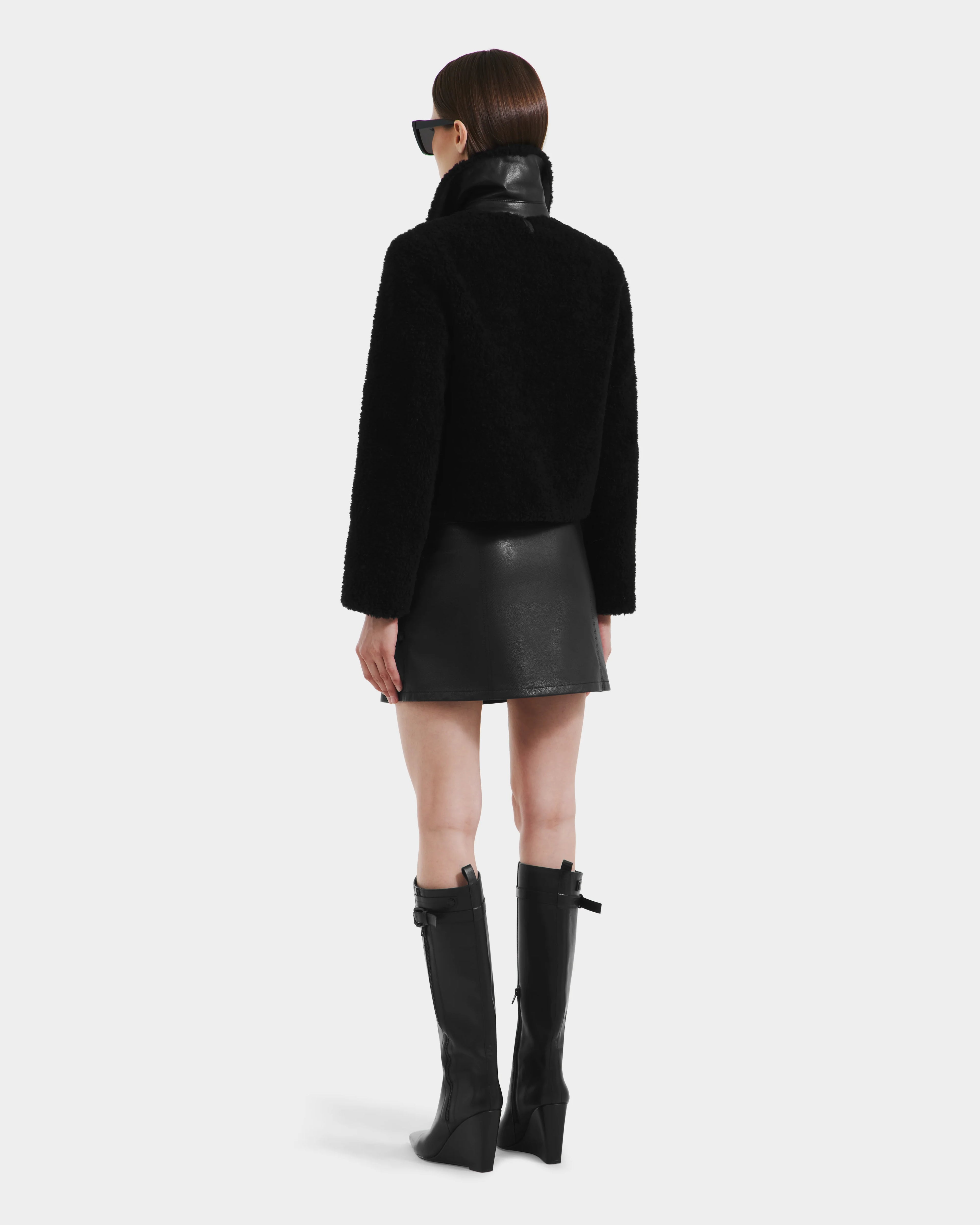 A woman with straight brown hair wears Rudsak’s BLAIR - 8125513 Black cropped teddy jacket, sunglasses, a black leather mini skirt, and black knee-high boots while facing away from the camera against a plain white background.