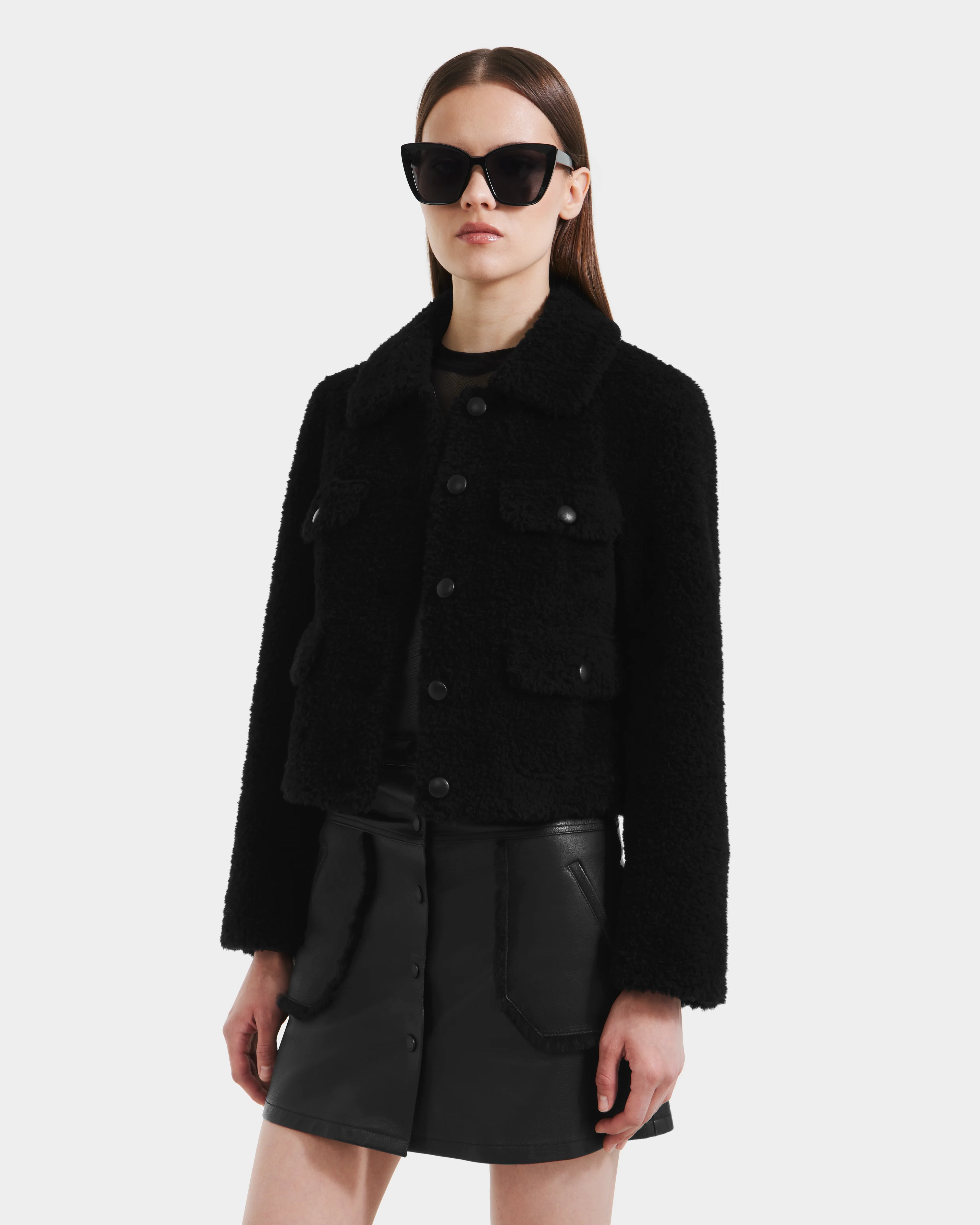 A woman wearing Rudsaks BLAIR - 8125513 Black modern cropped teddy jacket with flap pockets, large black sunglasses, and a black leather mini skirt stands against a plain light background.