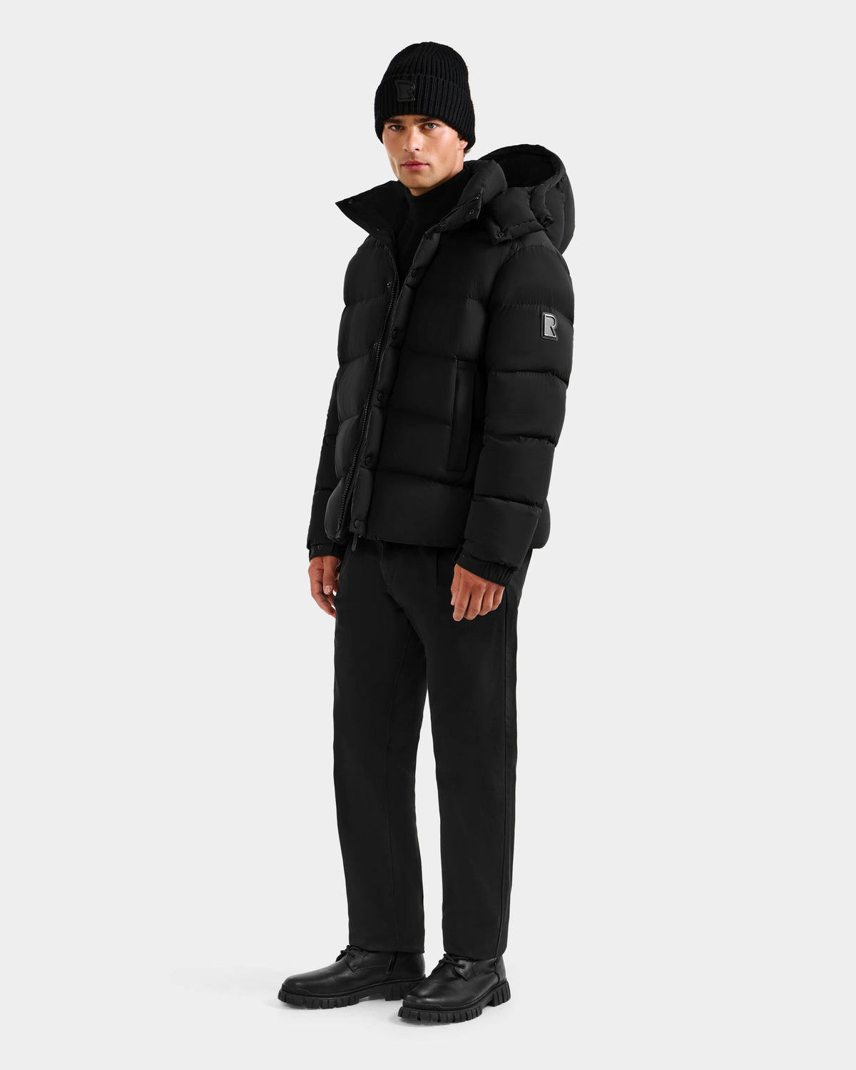 A man wearing the Rudsak BRAYDEN M-S - M125524 Black puffer jacket, black knit beanie, black pants, and black shoes stands against a plain light gray background.