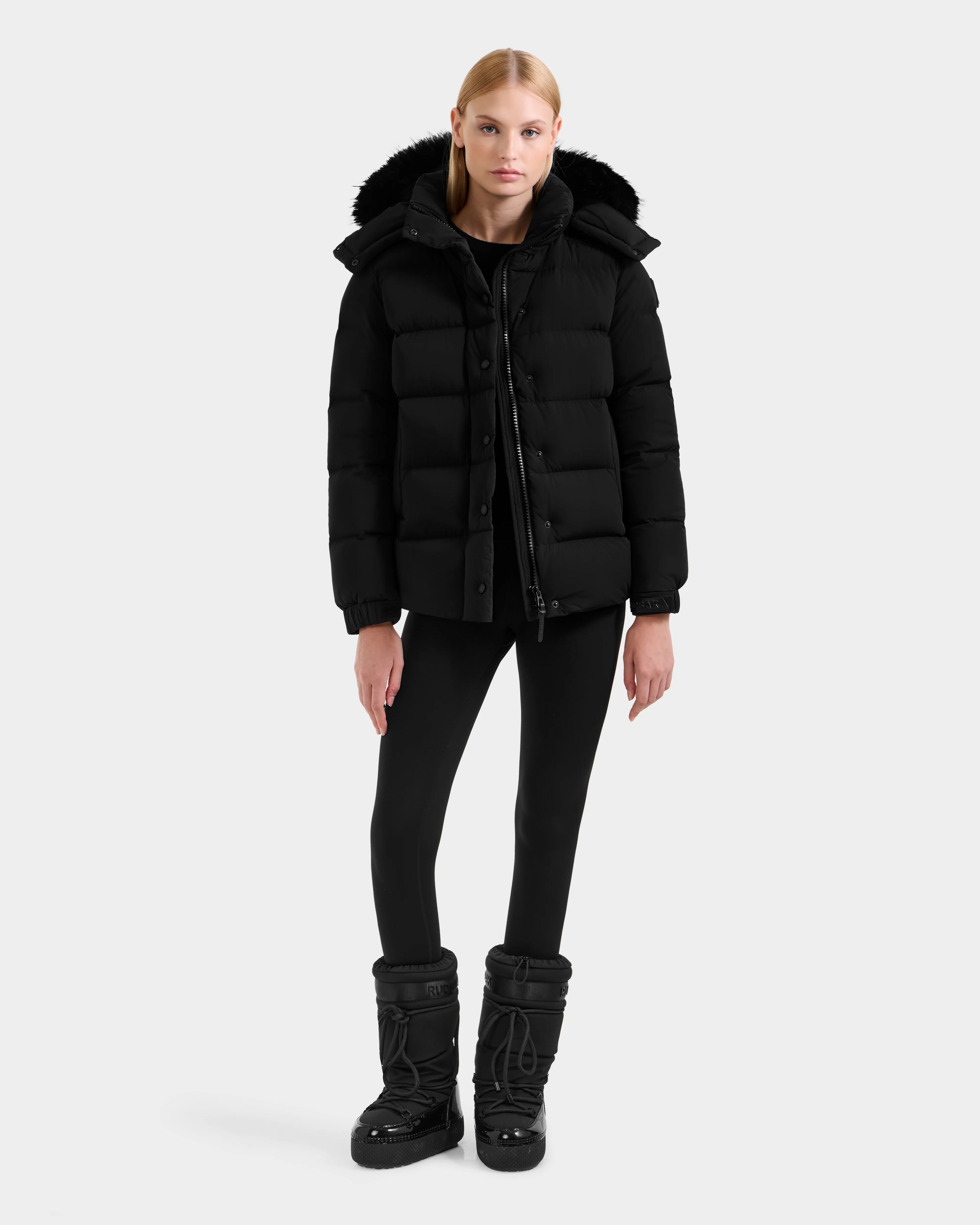 A person stands facing forward in the Rudsak BREE M - L125582 Black women’s down puffer jacket with a fur-trimmed removable hood, black leggings, and winter boots against a plain light background—a cold-weather essential.