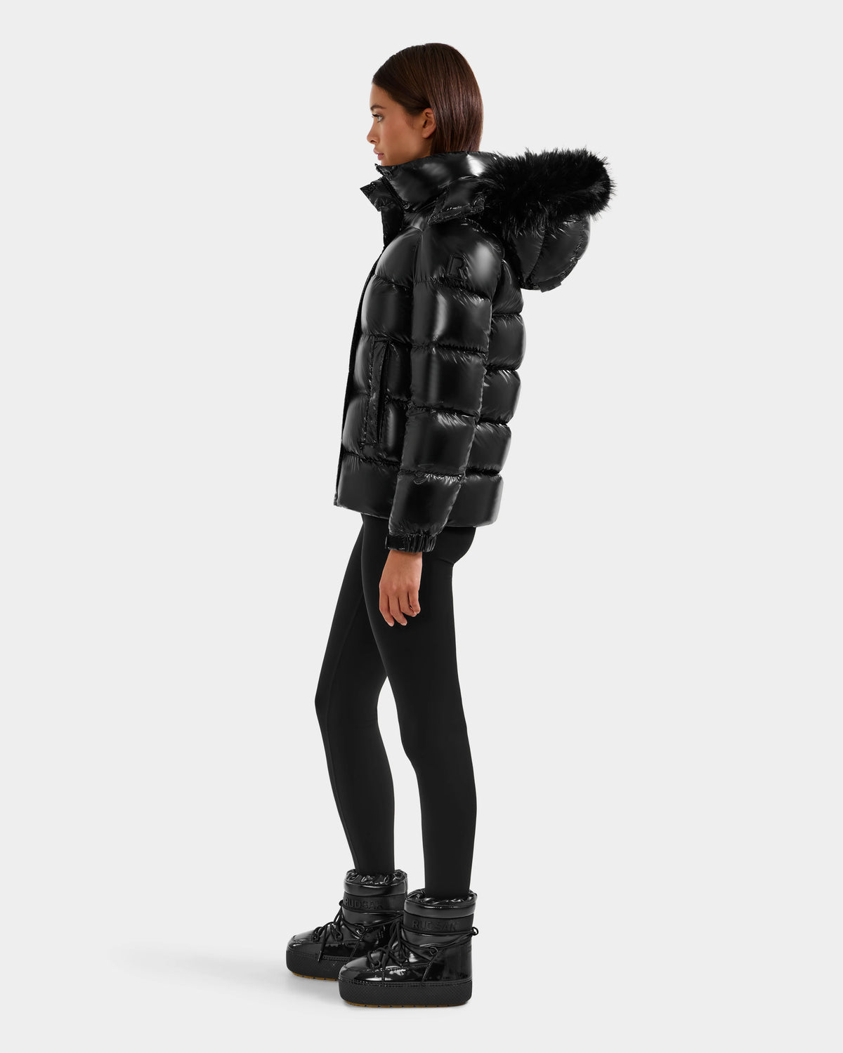 A woman in profile wears the Rudsak BREE S - L125583 Black down puffer jacket with a removable fur-lined hood, paired with black leggings and winter boots, standing against a plain light background.